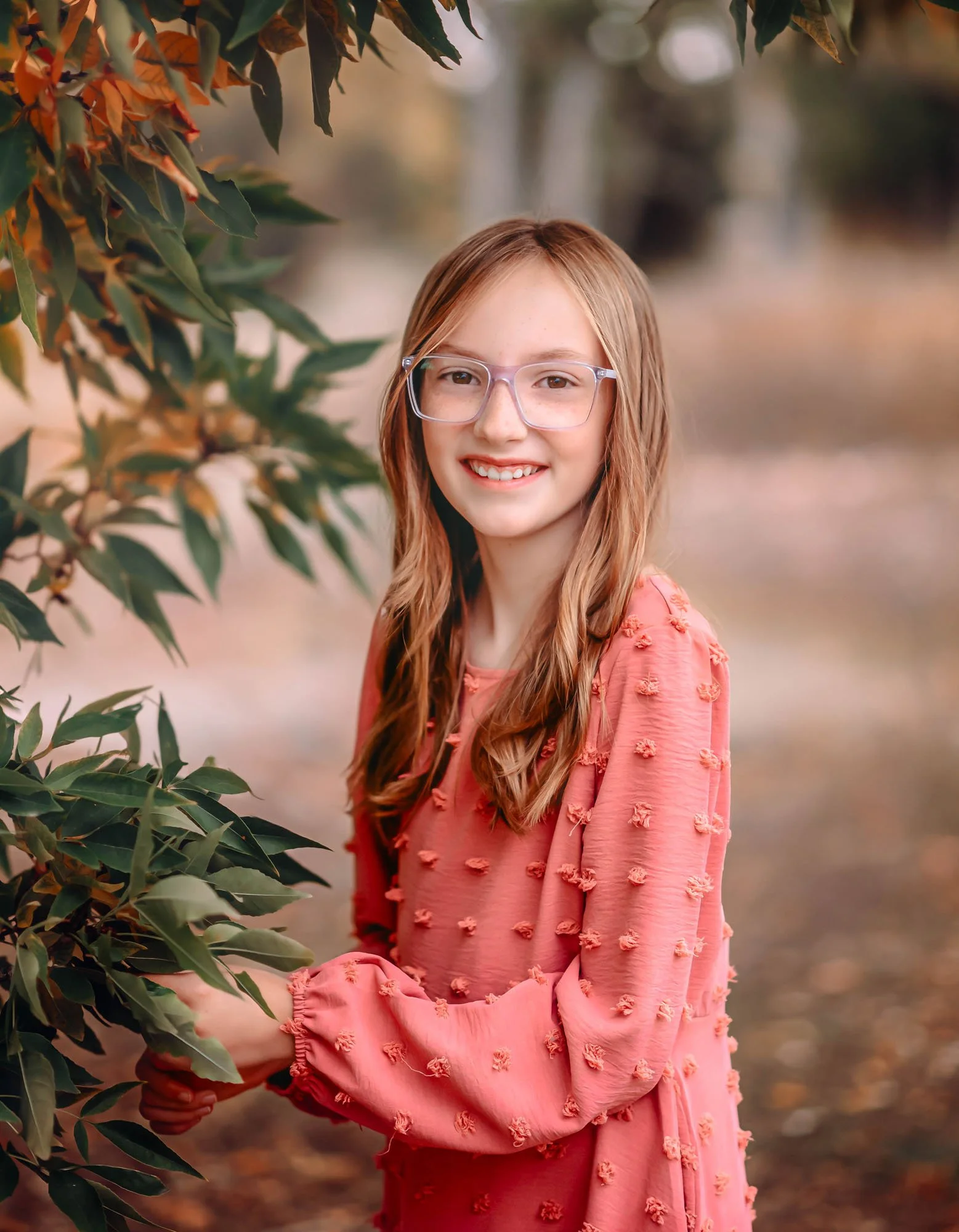 A young girl with long, wavy light brown hair and clear glasses, smiling outdoors in a pink dress with textured pom-pom details, surrounded by autumnal foliage.