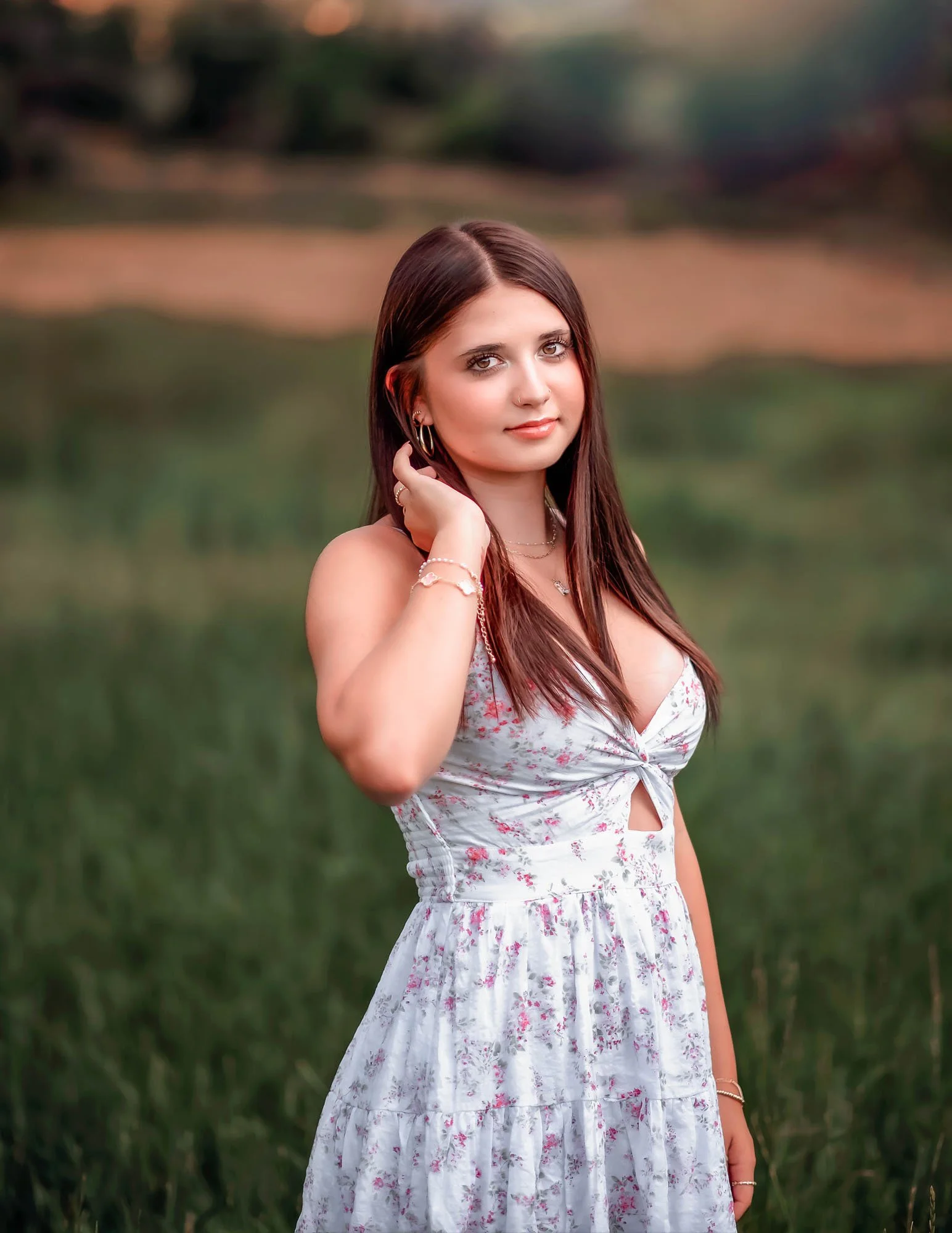 A young woman with long brown hair and a nose ring standing outdoors in a natural setting with green grass and blurred trees in the background. She is wearing a white dress with pink floral patterns and jewelry, with her hand touching her hair and lo