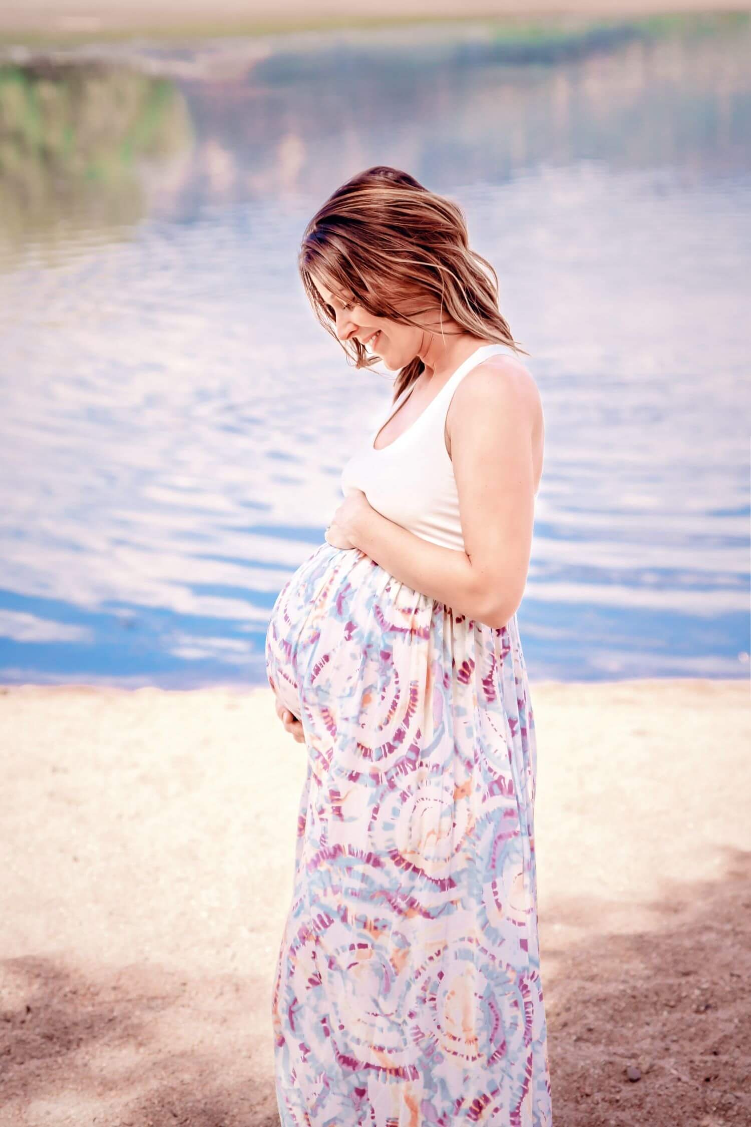 A pregnant woman standing on a beach with her head bowed and a gentle smile, wearing a white top and a colorful, patterned maxi skirt, with a calm body of water in the background.