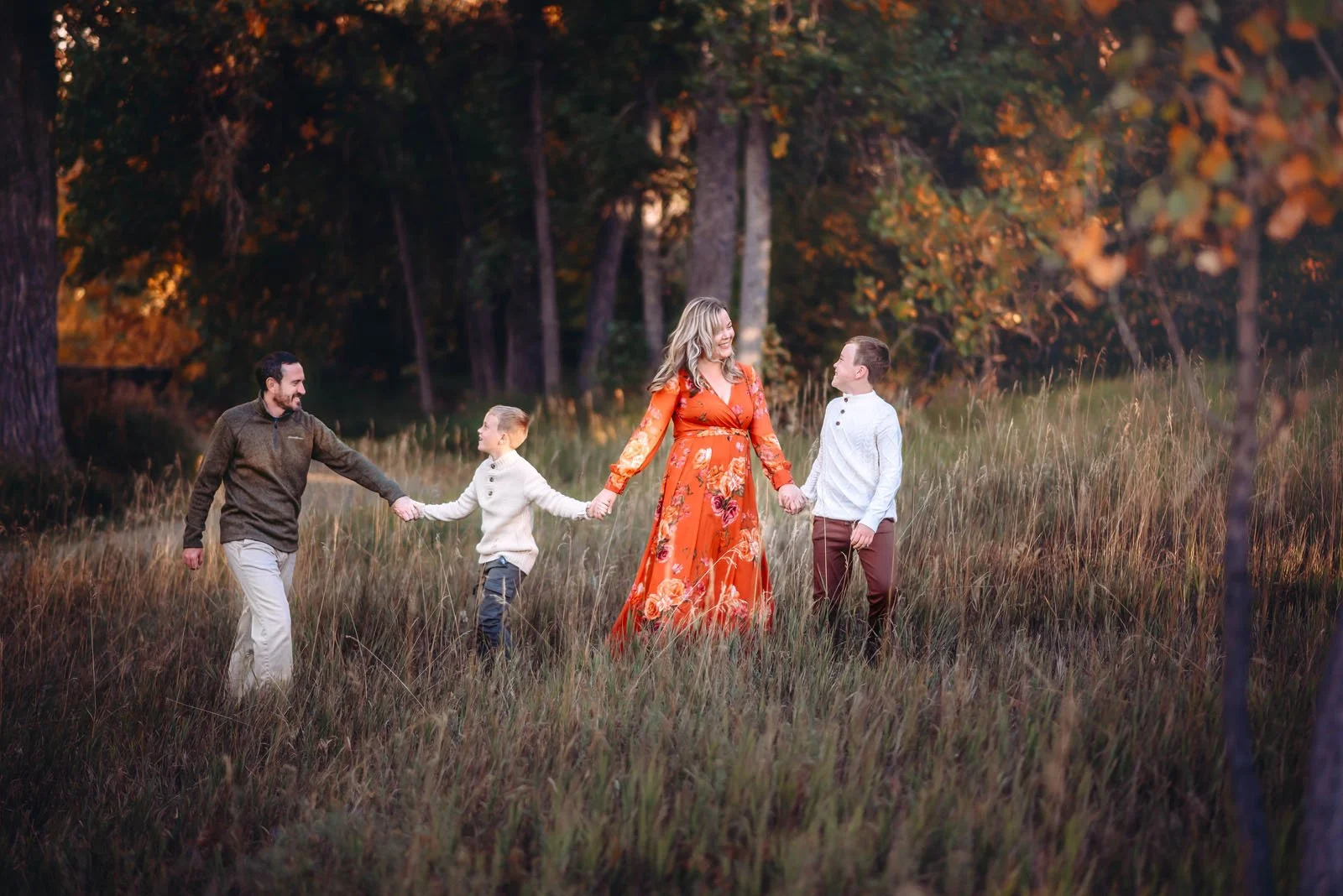 A family of four holding hands and walking through a field of tall grass in a forest during autumn. The mother, wearing an orange floral dress, is in the center, with a father on her left and two young boys on her right and left, wearing sweaters.