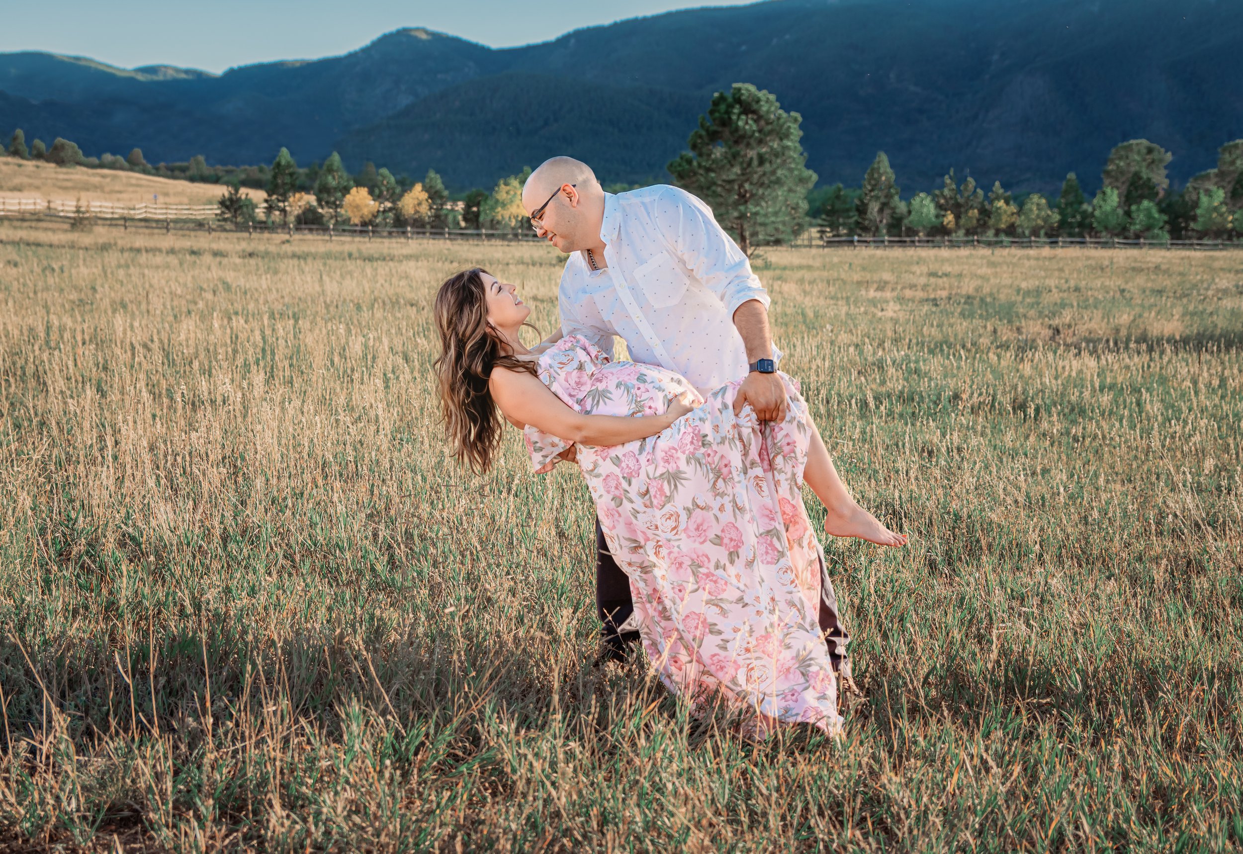 A man is holding a woman in a field of tall grass with mountains and trees in the background, during the daytime.