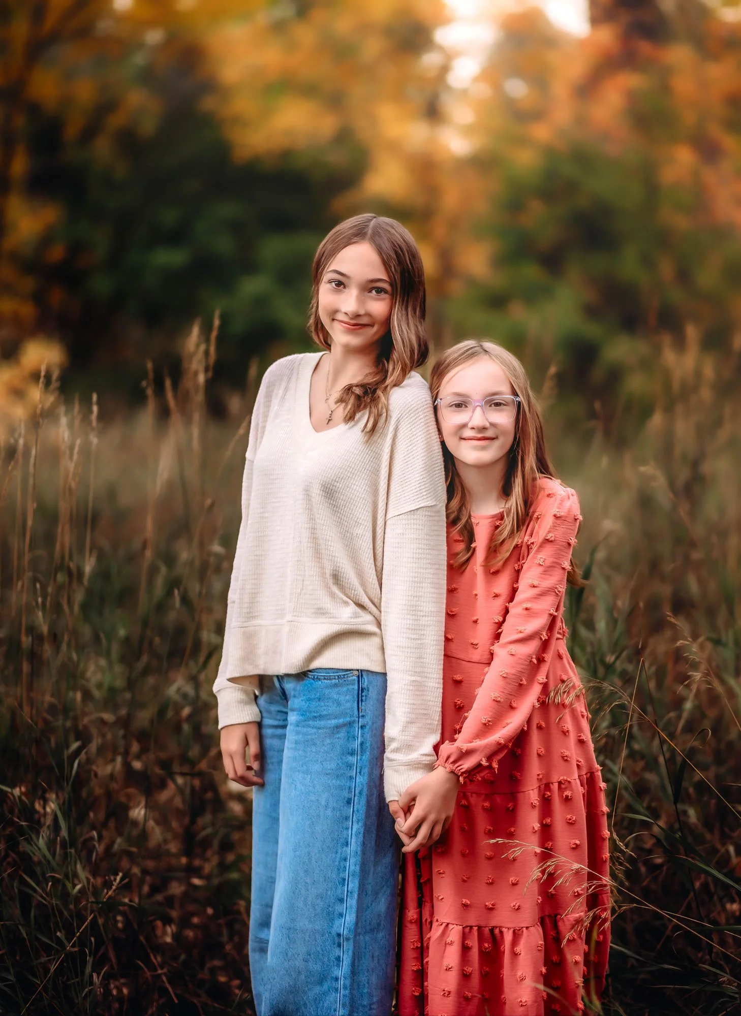 Two girls standing outdoors in a field with orange and green autumn leaves in the background. The girl on the left has light brown hair, is wearing a cream sweater and blue jeans. The girl on the right has long hair, glasses, and is wearing an orange