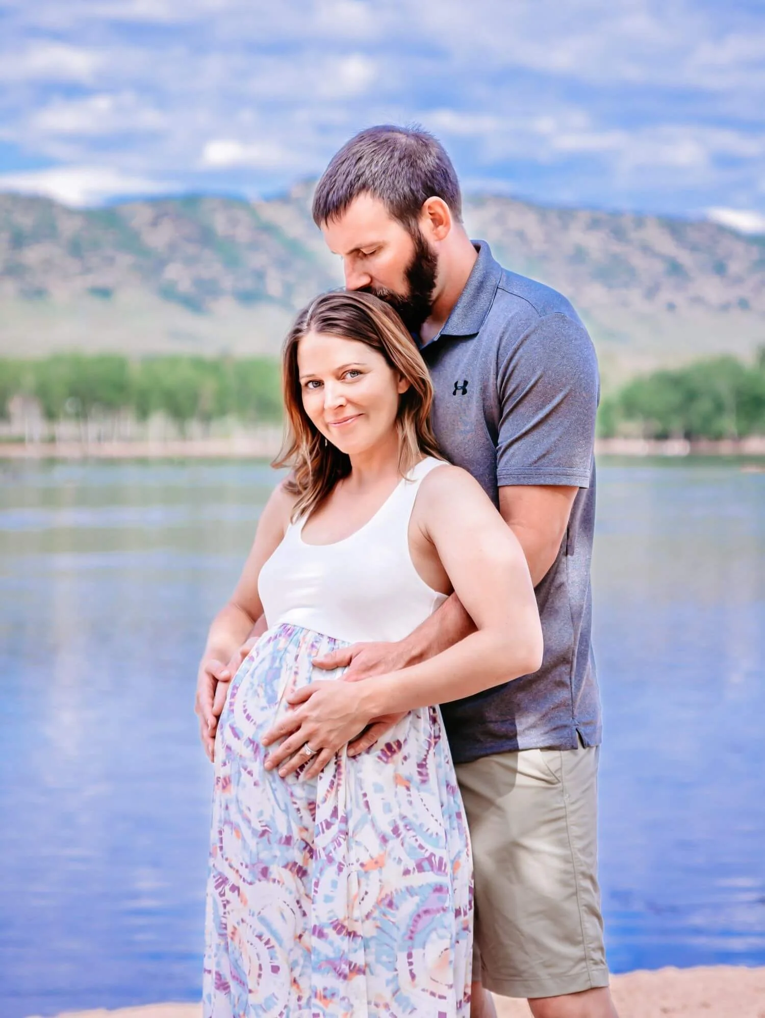 A pregnant woman in a white and pastel-colored dress stands by a body of water with a mountain backdrop, while a man in a blue polo shirt and beige shorts embraces her from behind, resting his head on her forehead.