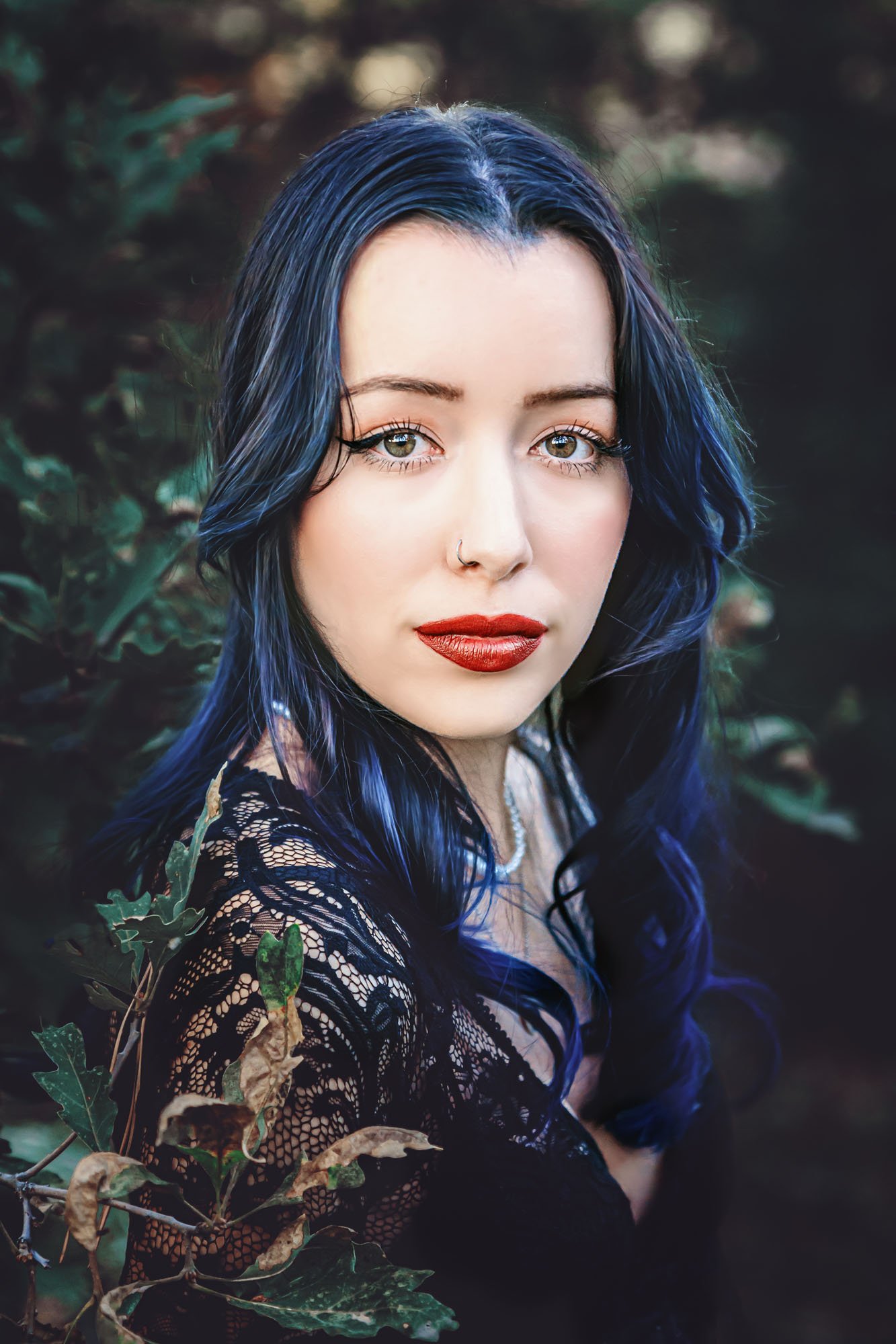 A young woman with blue hair and hazel eyes looking at the camera in an outdoor setting with dark foliage in the background.