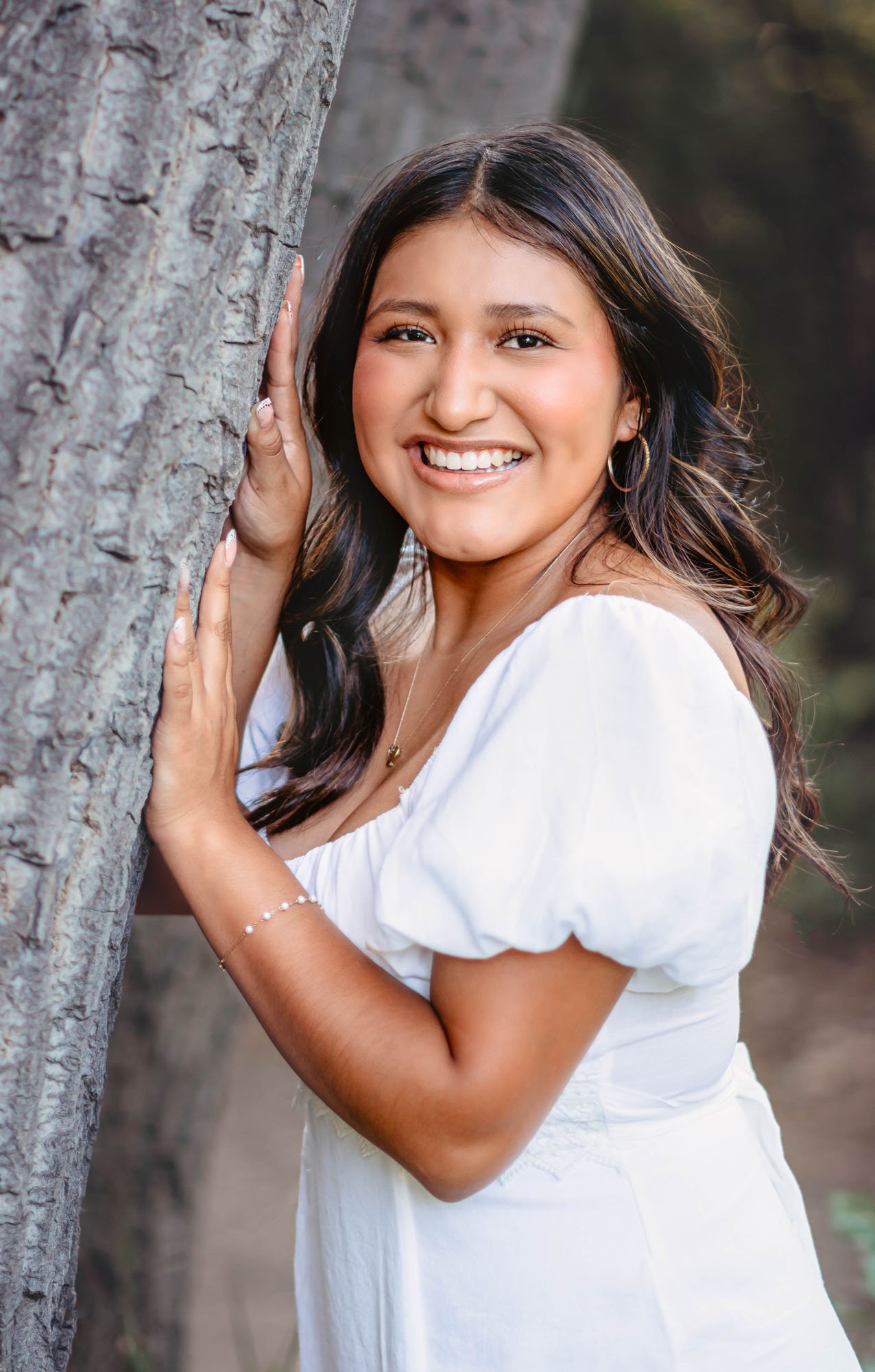 A woman with long dark hair smiling and wearing a white dress, standing outdoors and leaning against a tree.