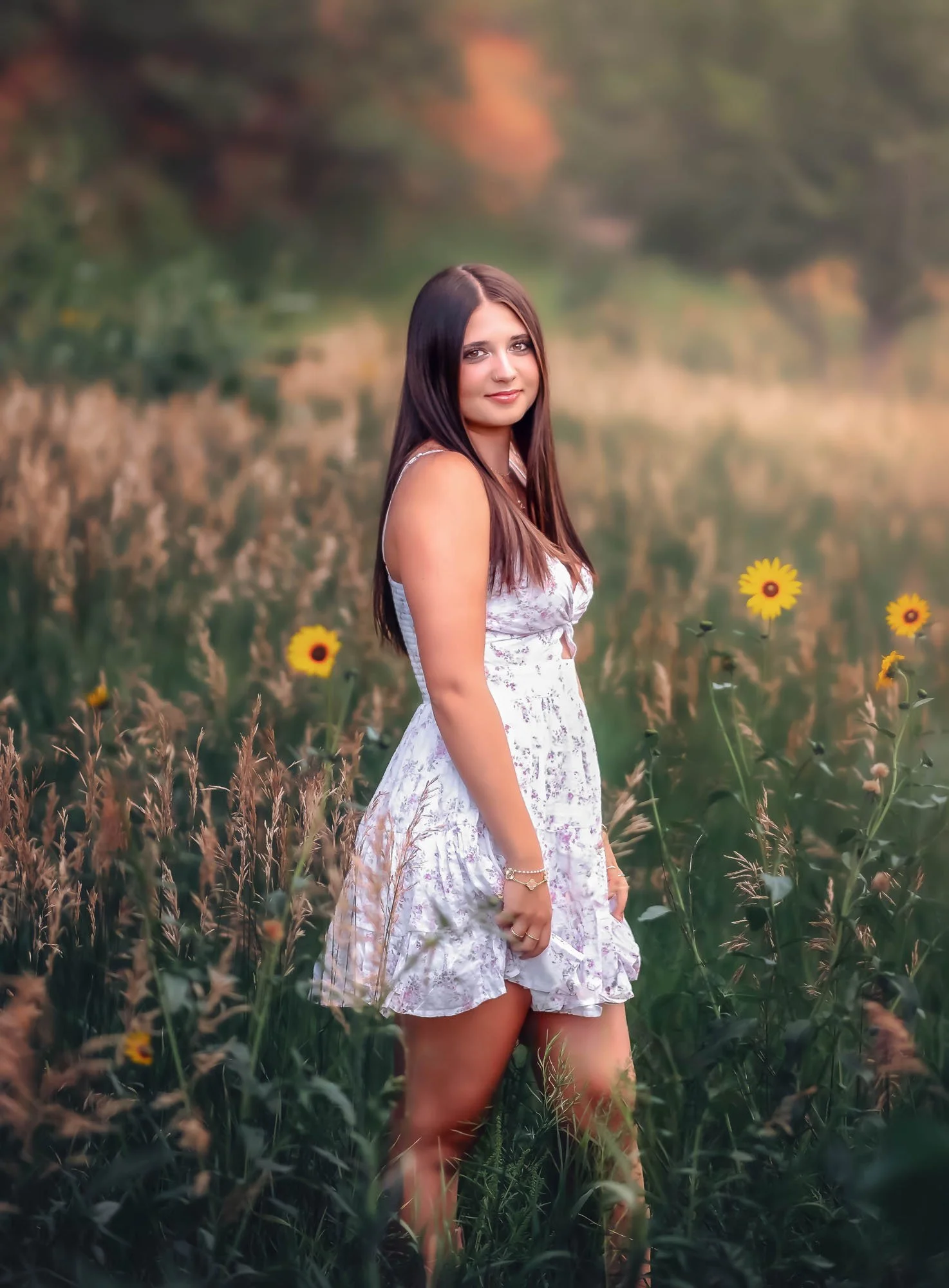 A young woman with long dark hair wearing a white floral dress standing in a field of tall grass and yellow flowers, with trees and a blurred background.