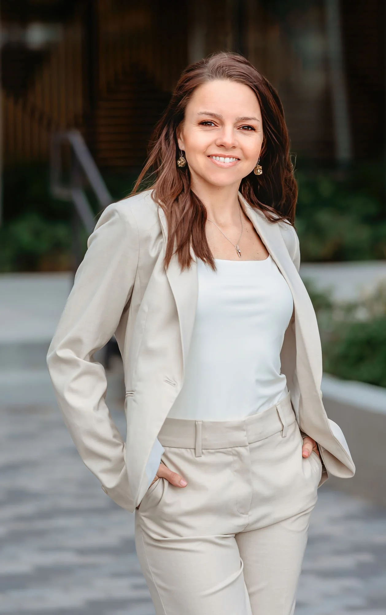 A woman with brown hair wearing a light-colored blazer and matching trousers, smiling outdoors with hands in pockets.