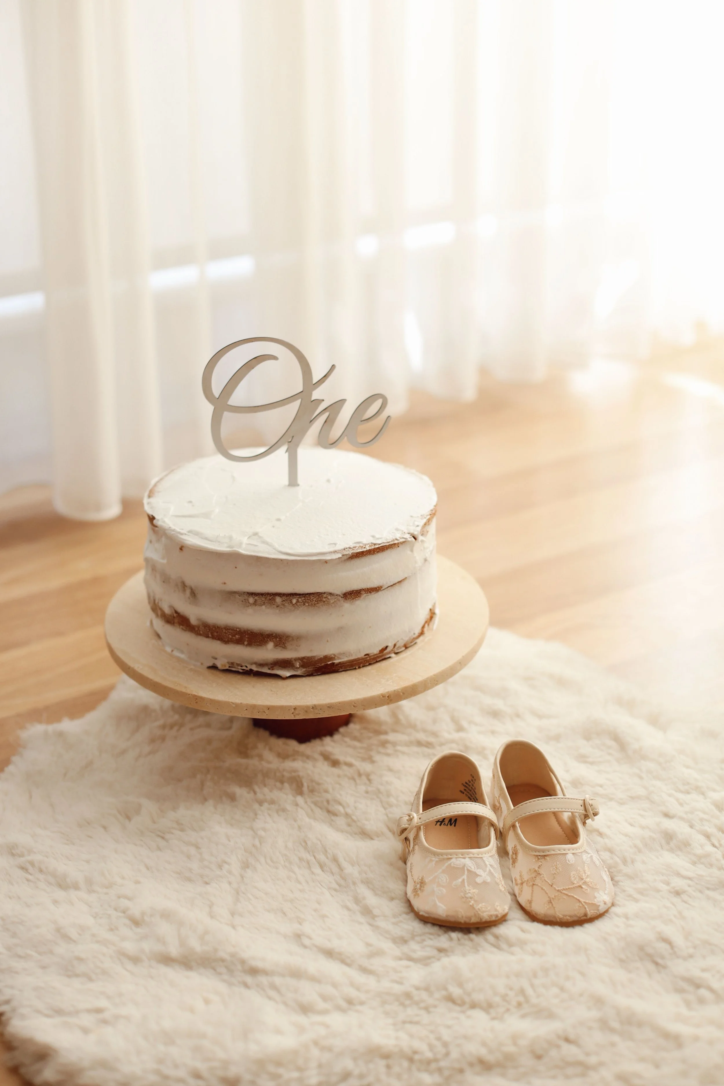 A small, round cake with white icing and a silver 'One' cake topper on a light wooden cake stand, set on a fluffy white rug in front of sheer white curtains. Nearby, white baby shoes with lace detail.