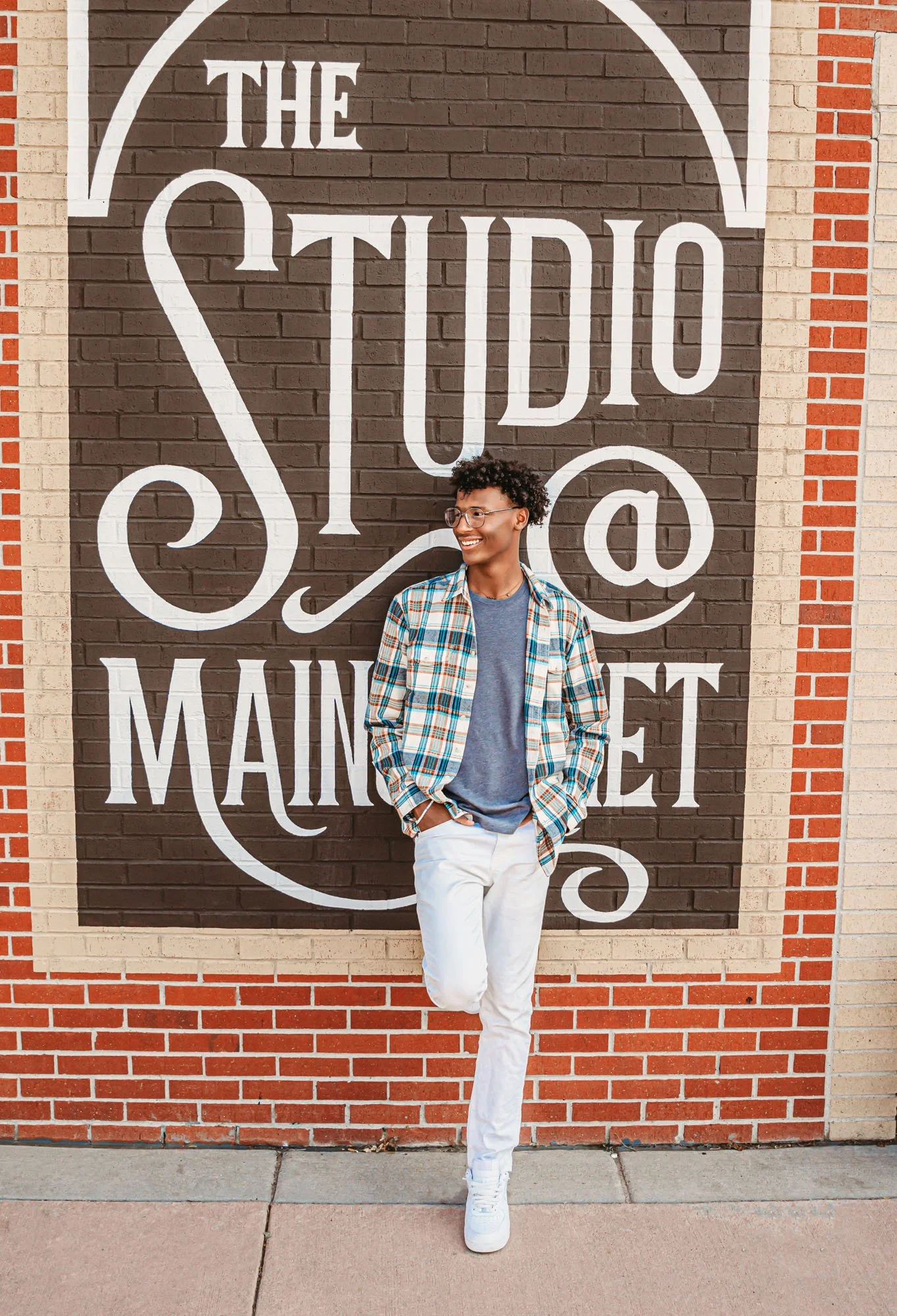 A young man with curly hair and glasses standing against a brick wall with a logo reading 'The Studio' in large white letters.
