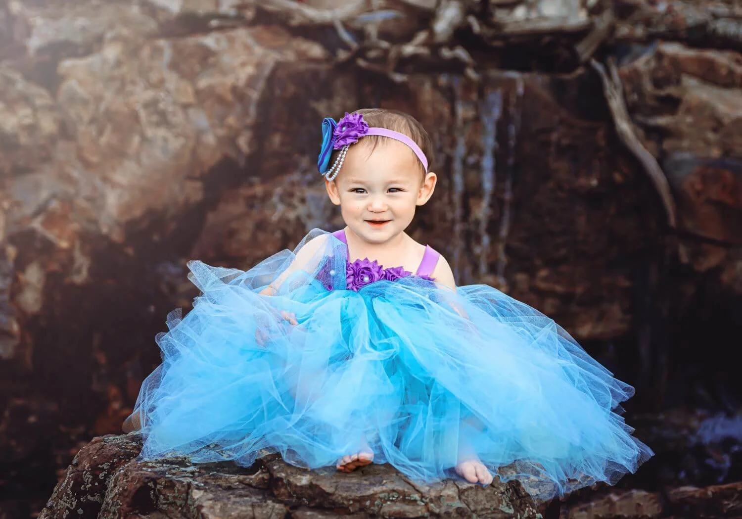 A young girl in a purple and blue princess dress with a tulle skirt, sitting on a rocky surface in front of a waterfall.