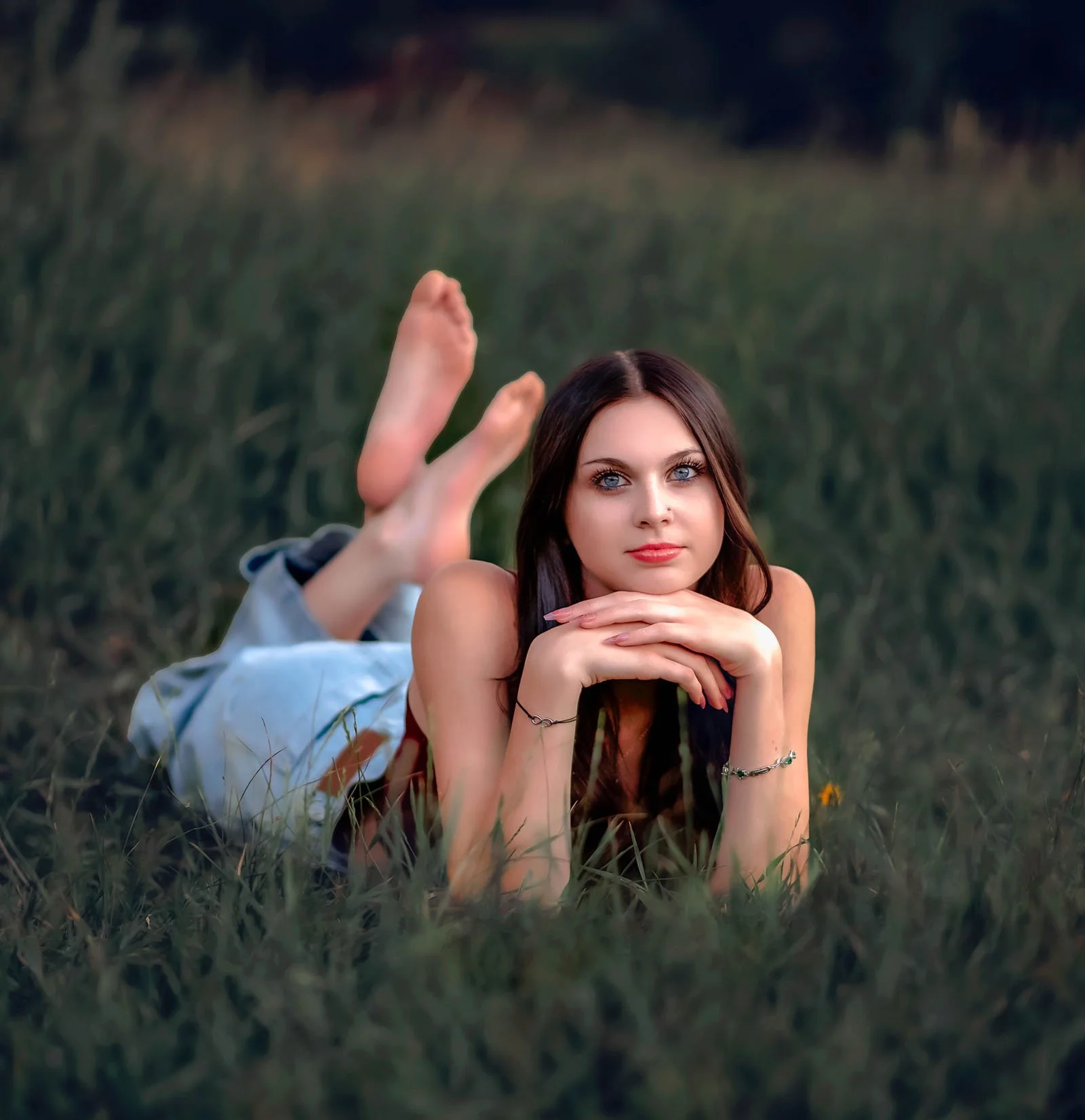 A young woman with blue eyes and long brown hair lying on her stomach in a grassy field, resting her chin on her folded arms, and looking directly at the camera. A person behind her is partially visible, with just their feet and part of their leg in 
