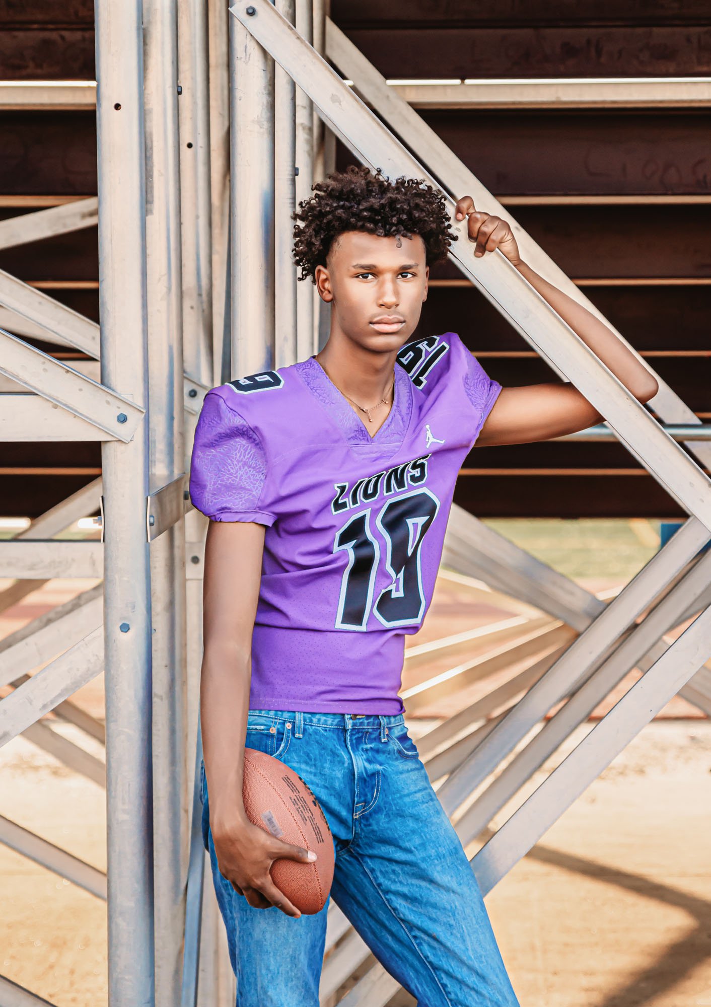 A teenage boy standing on metal bleachers holding a football, wearing a purple football jersey and blue jeans, with short curly hair, in an outdoor setting.