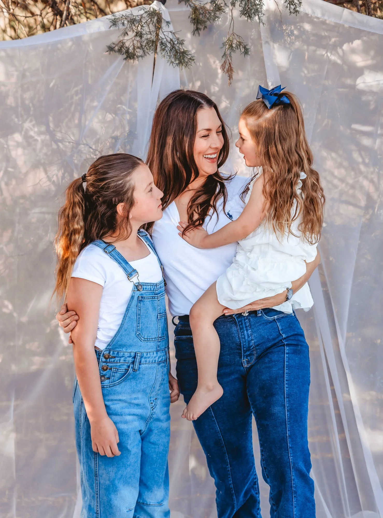A woman with two young girls, one of whom she is holding, all smiling and facing each other. The woman and the girl she is holding are wearing white shirts and blue jeans, while the other girl wears a white shirt and denim overalls. They are outdoors