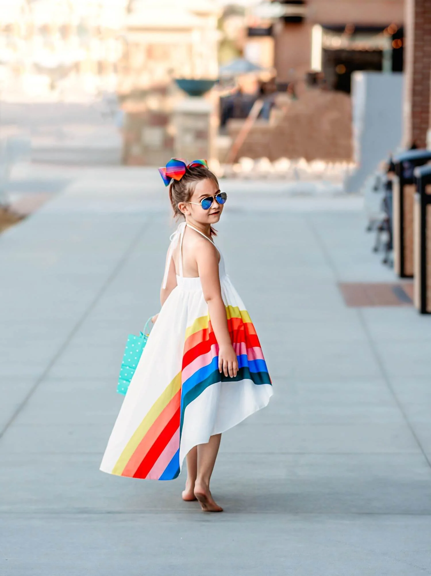 A young girl walking barefoot on a sidewalk, wearing sunglasses, a white dress with rainbow stripes, and a large rainbow bow in her hair, carrying a polka dot bag.