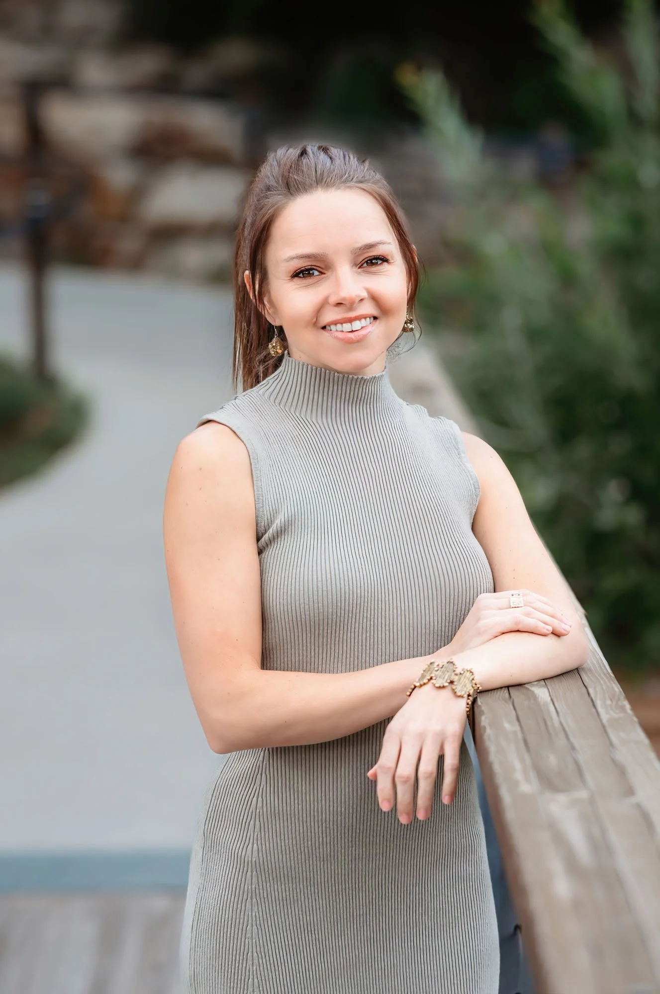 A smiling woman with brown hair and earrings leaning on a wooden railing outdoors, with a blurred natural background.