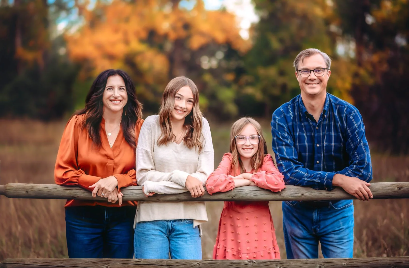 A family of five members standing outdoors on a wooden bridge with autumn trees in the background, smiling for the camera.