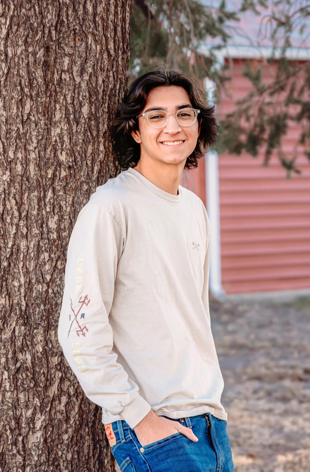 A young man with wavy dark hair and glasses leaning against a large tree, smiling at the camera outdoors, with blurred trees and a building in the background.
