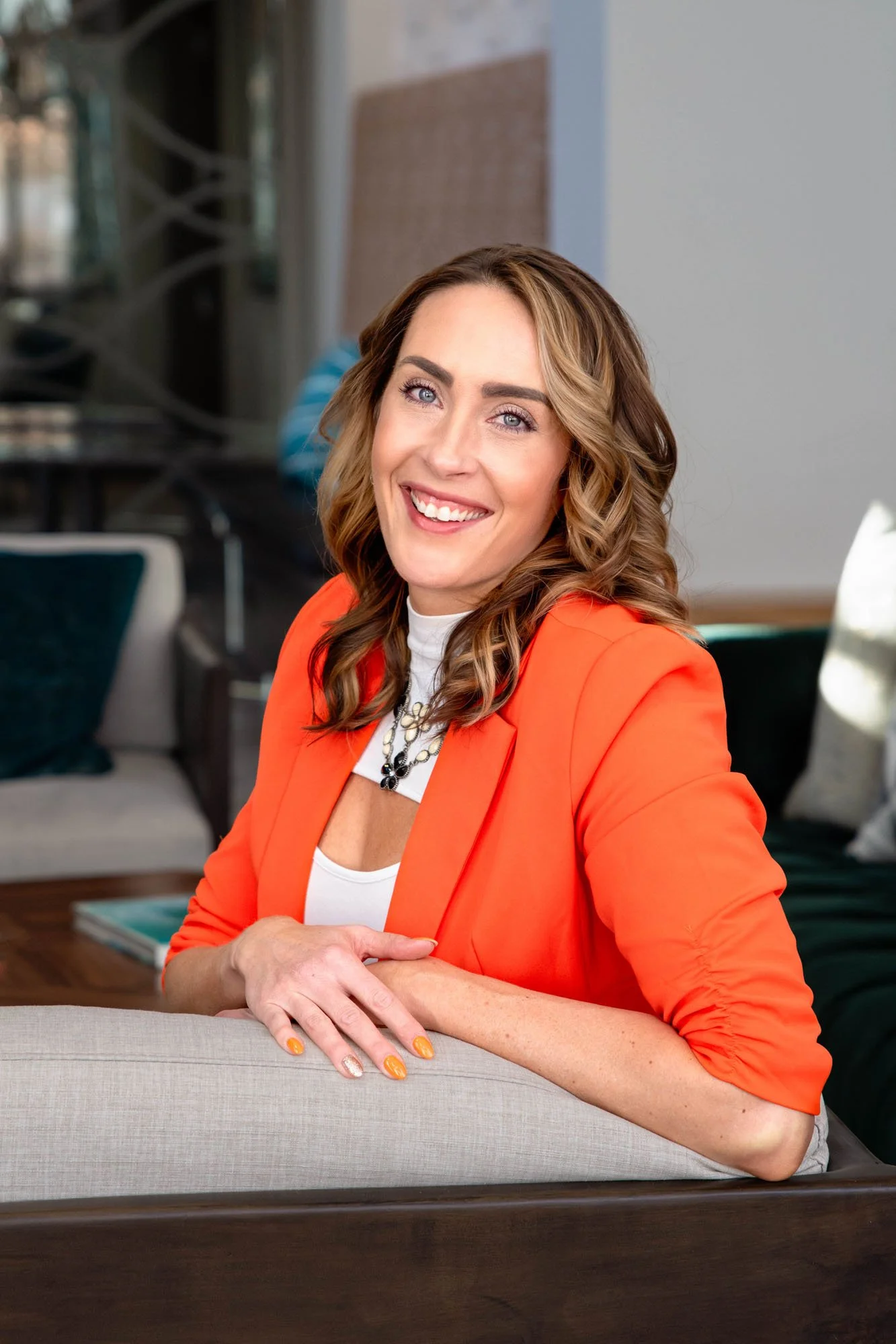 A smiling woman with brown curly hair and blue eyes, wearing an orange blazer and a necklace, is sitting at a table in a modern room.