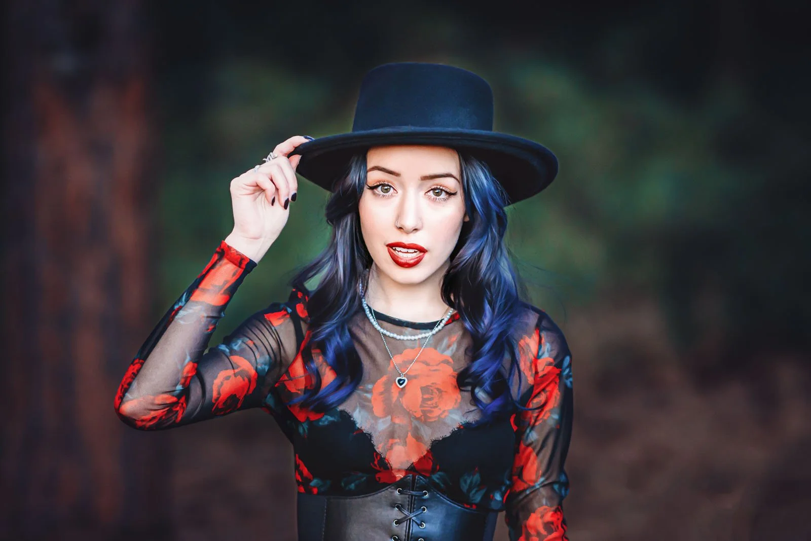 A woman with blue hair wearing a black wide-brimmed hat, a red floral sheer top, and layered necklaces, standing outdoors with a blurred background of trees.