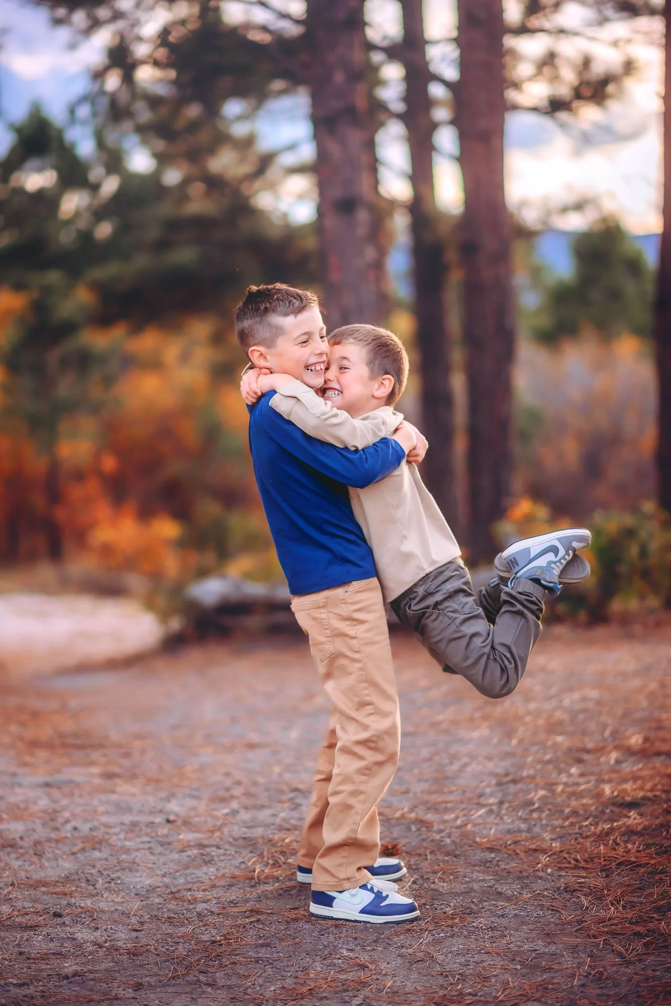 Two young boys with short hair embracing and playing cheerfully outdoors in a wooded park during autumn, with trees and fallen leaves in the background.