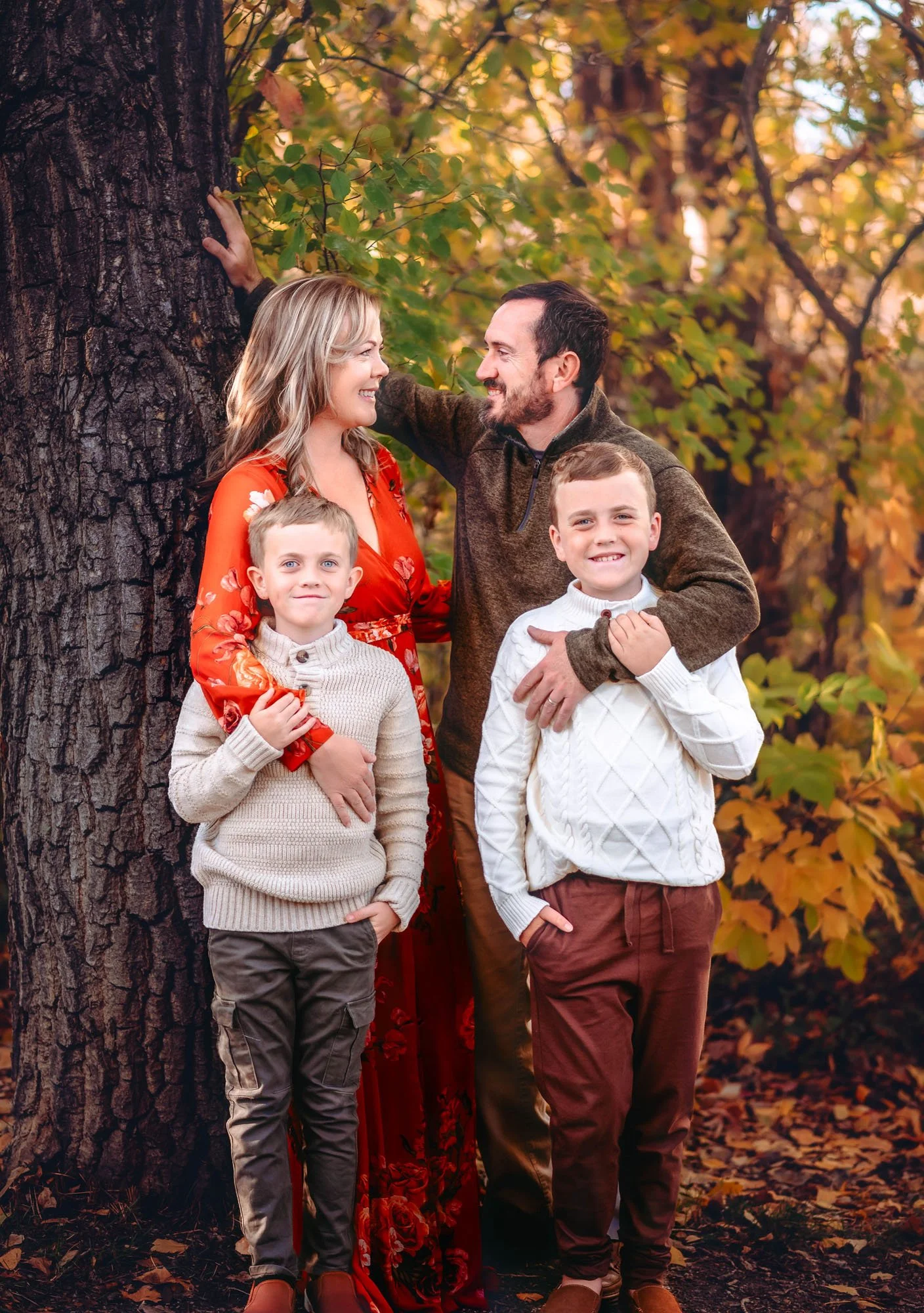 A family of four, including two young boys, a woman in a red dress, and a man, posing outdoors during autumn. They are standing next to a large tree with fall foliage in the background, and fallen leaves on the ground.