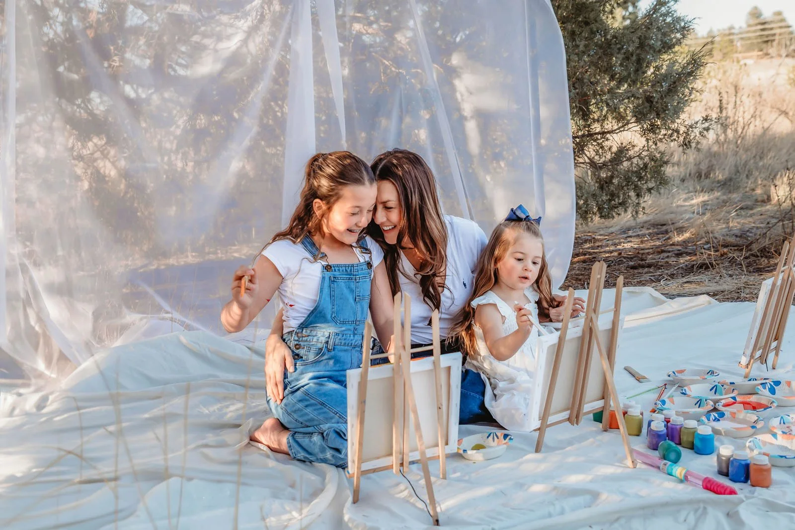 A woman and two young girls painting on small canvases outdoors on a white sheet with painting supplies nearby, surrounded by trees and natural scenery.