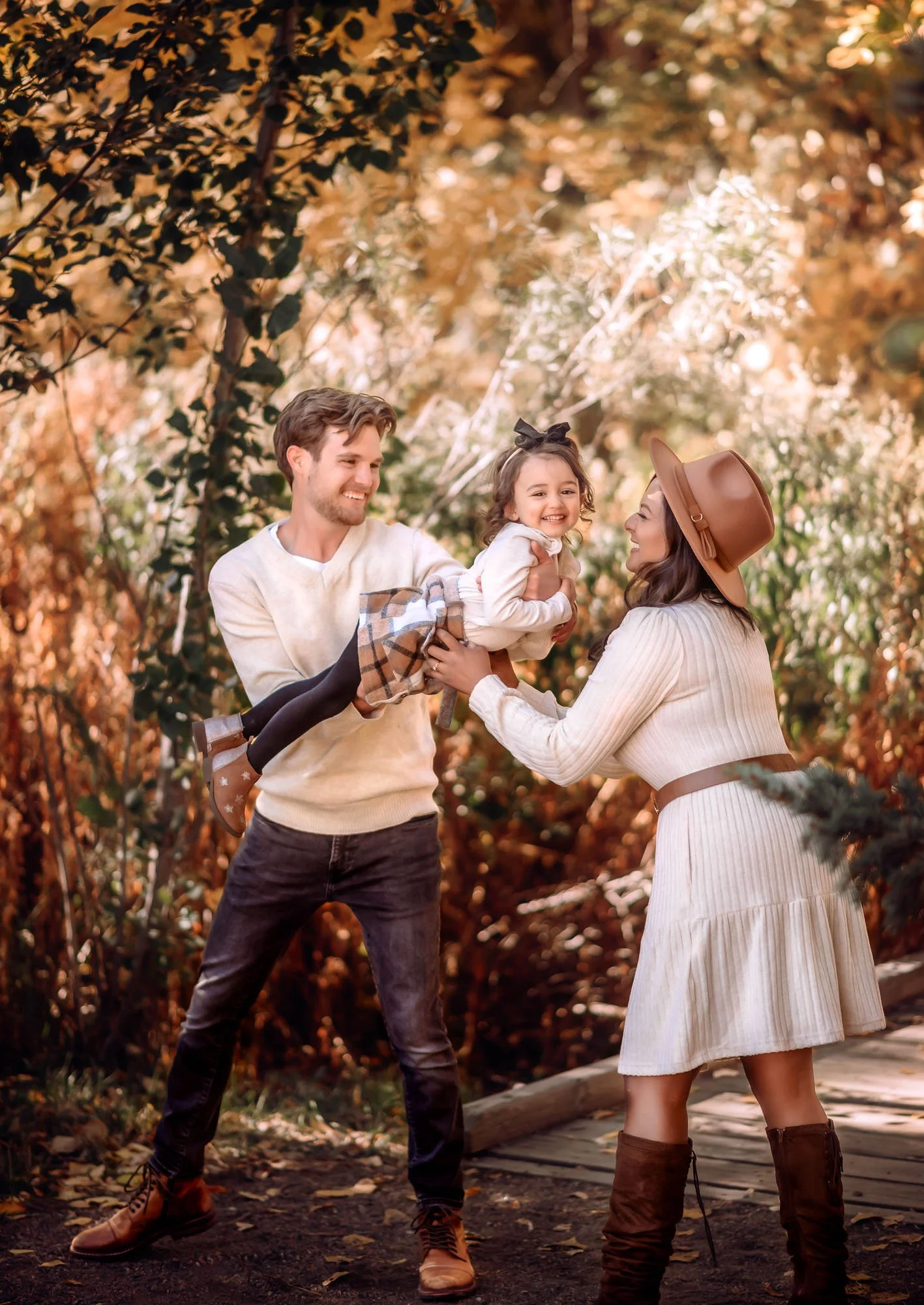 A family of three enjoying fall outdoors, with the father holding a young girl and the mother smiling at her, surrounded by autumn leaves.