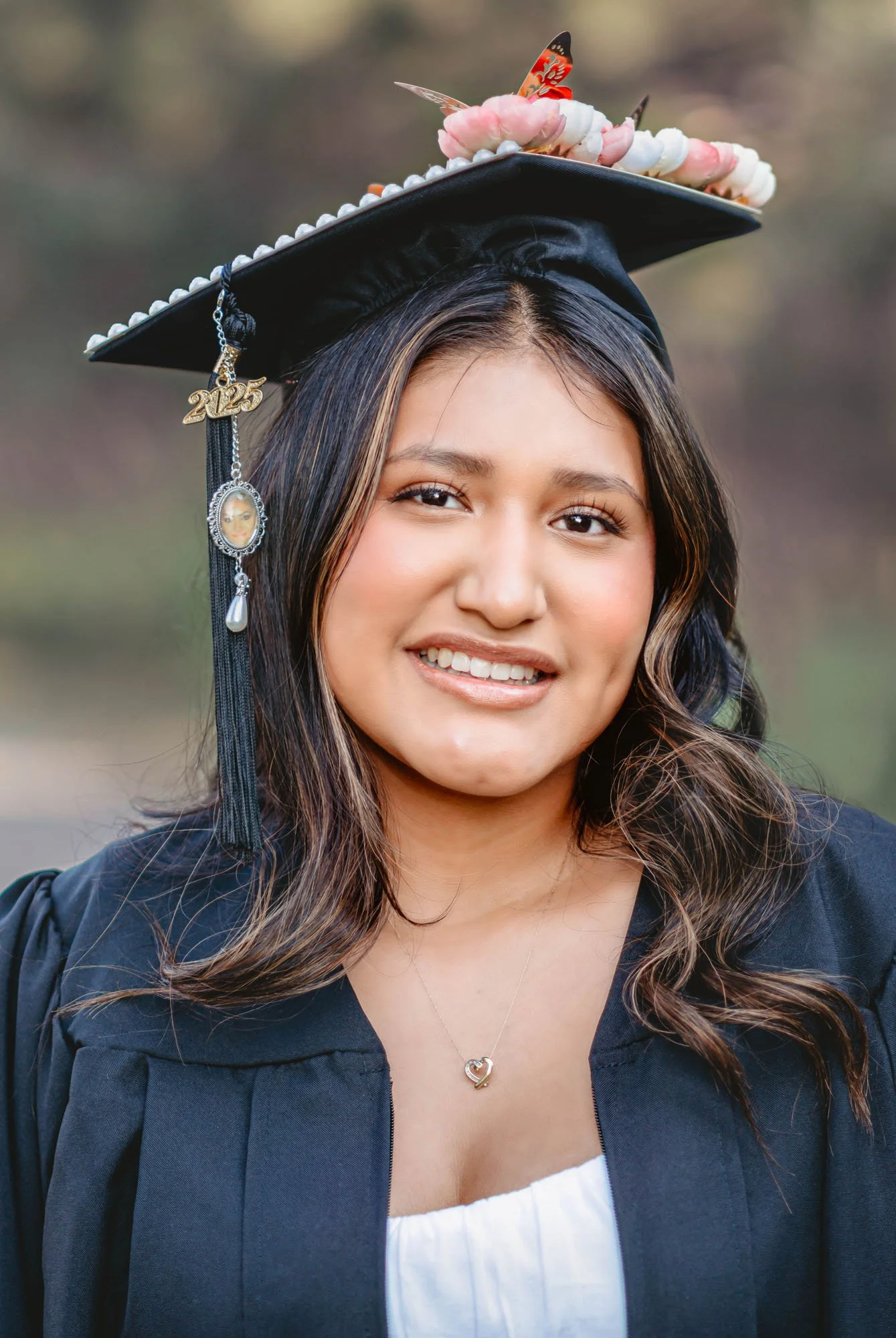 Young woman in graduation cap and gown smiling, outdoors, wearing a necklace with a heart-shaped pendant.