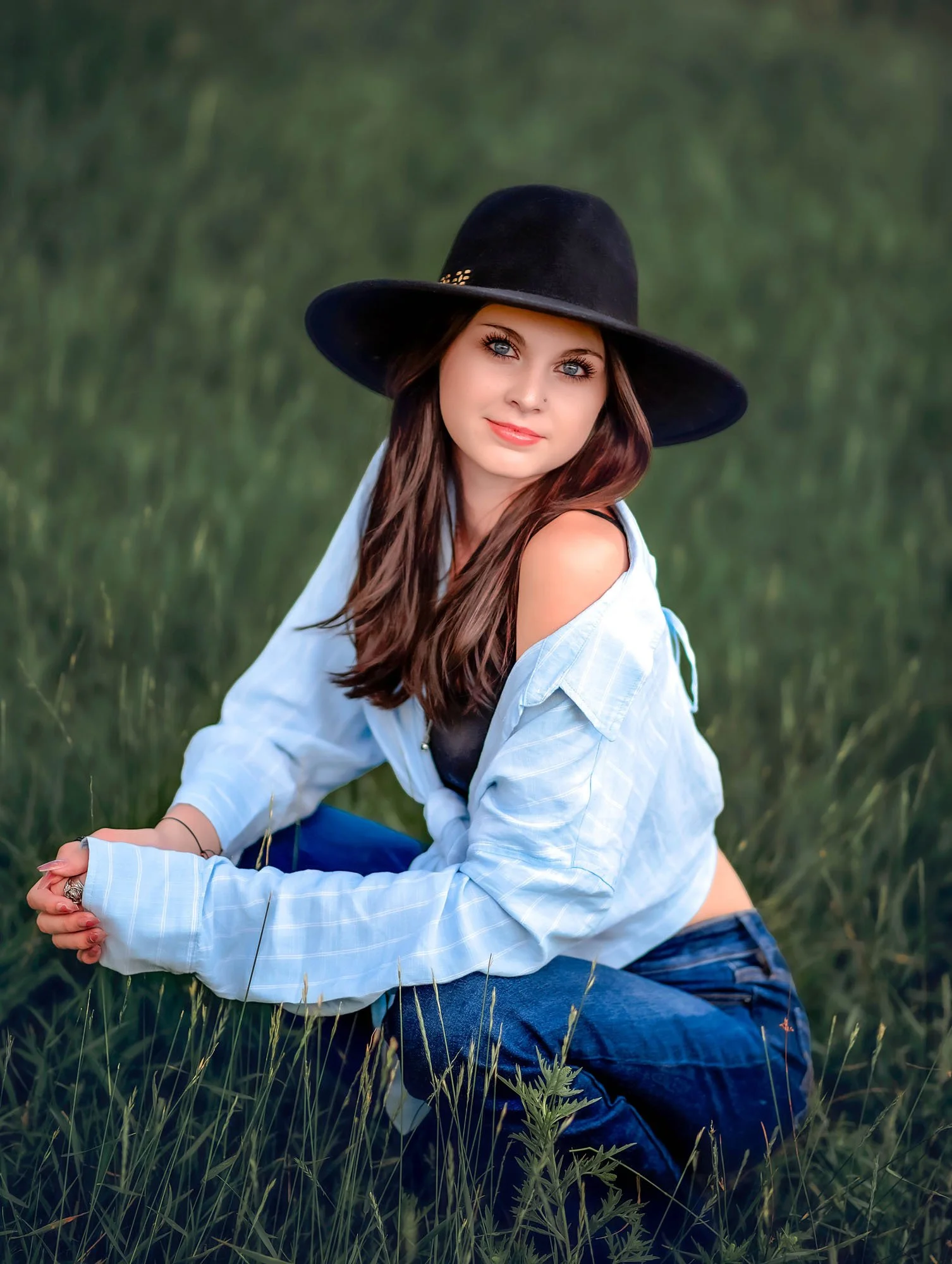 A young woman with long brown hair and blue eyes sitting in a grassy field, wearing a black wide-brimmed hat, a white shirt, and blue jeans.
