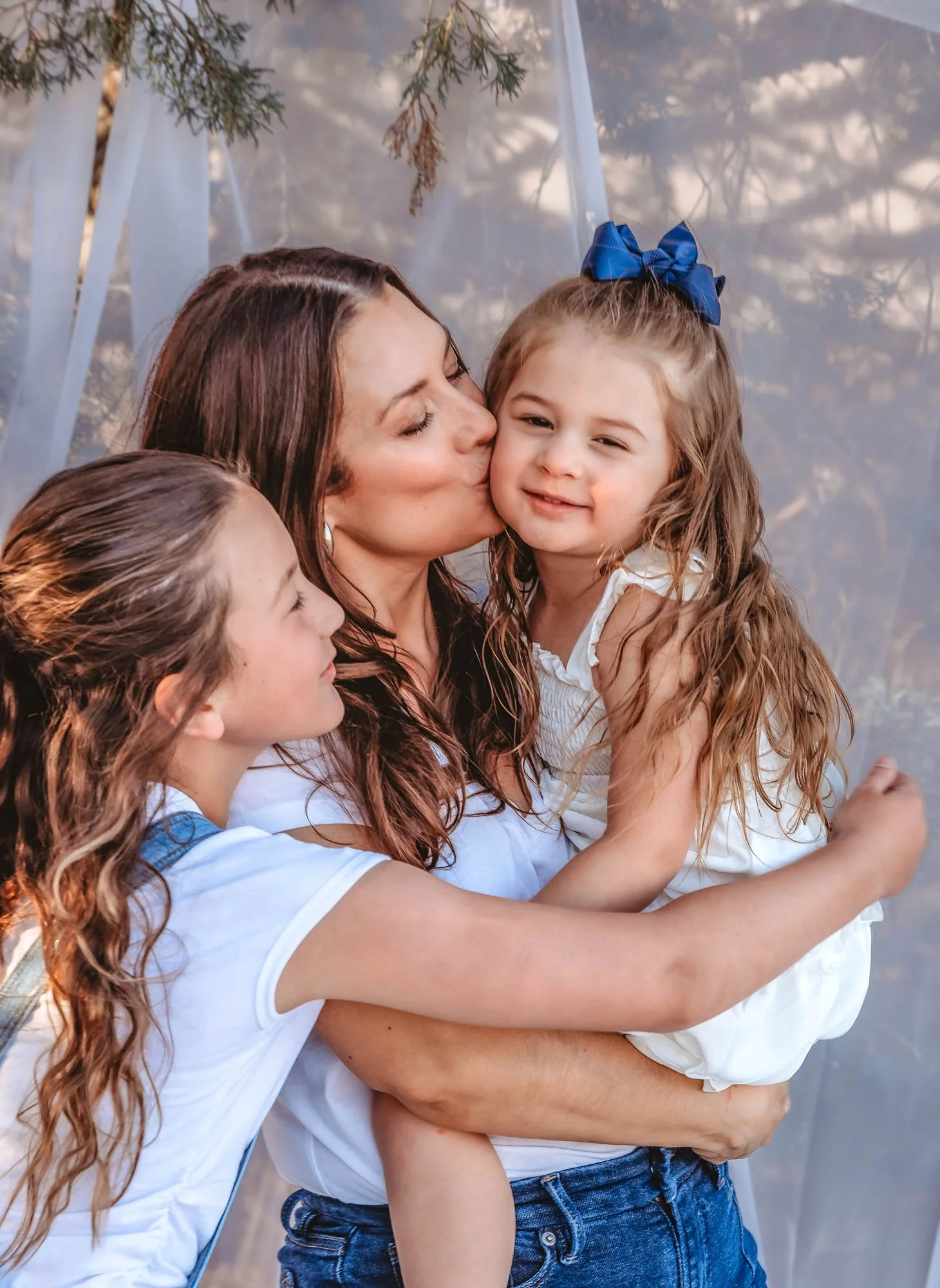 A woman hugging and kissing a young girl with long, curly hair and a large blue bow in her hair, while another girl with long wavy hair looks on.