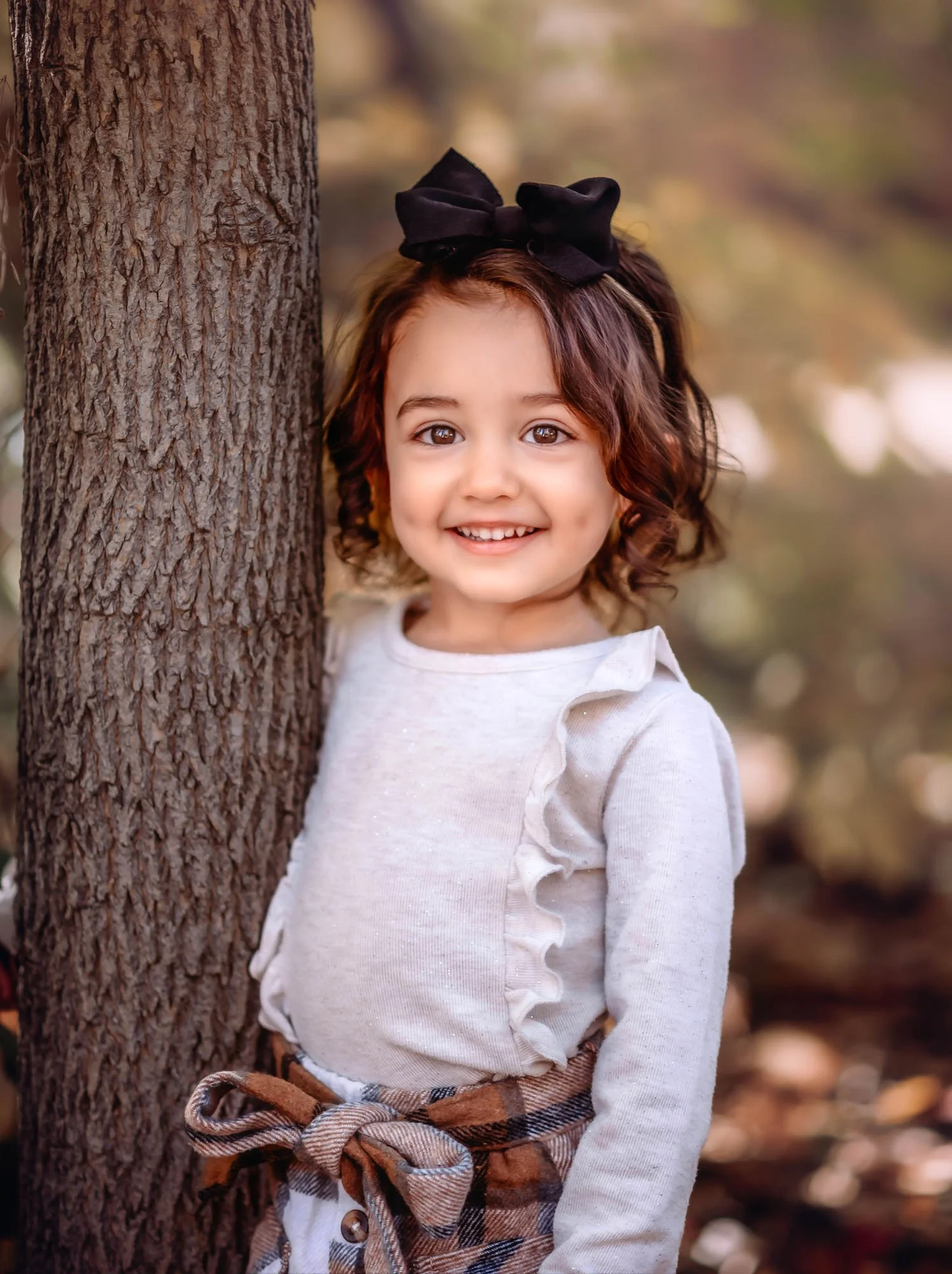 A young girl with brown curly hair and a black bow headband standing outdoors next to a tree, smiling at the camera, with a blurred autumn background.