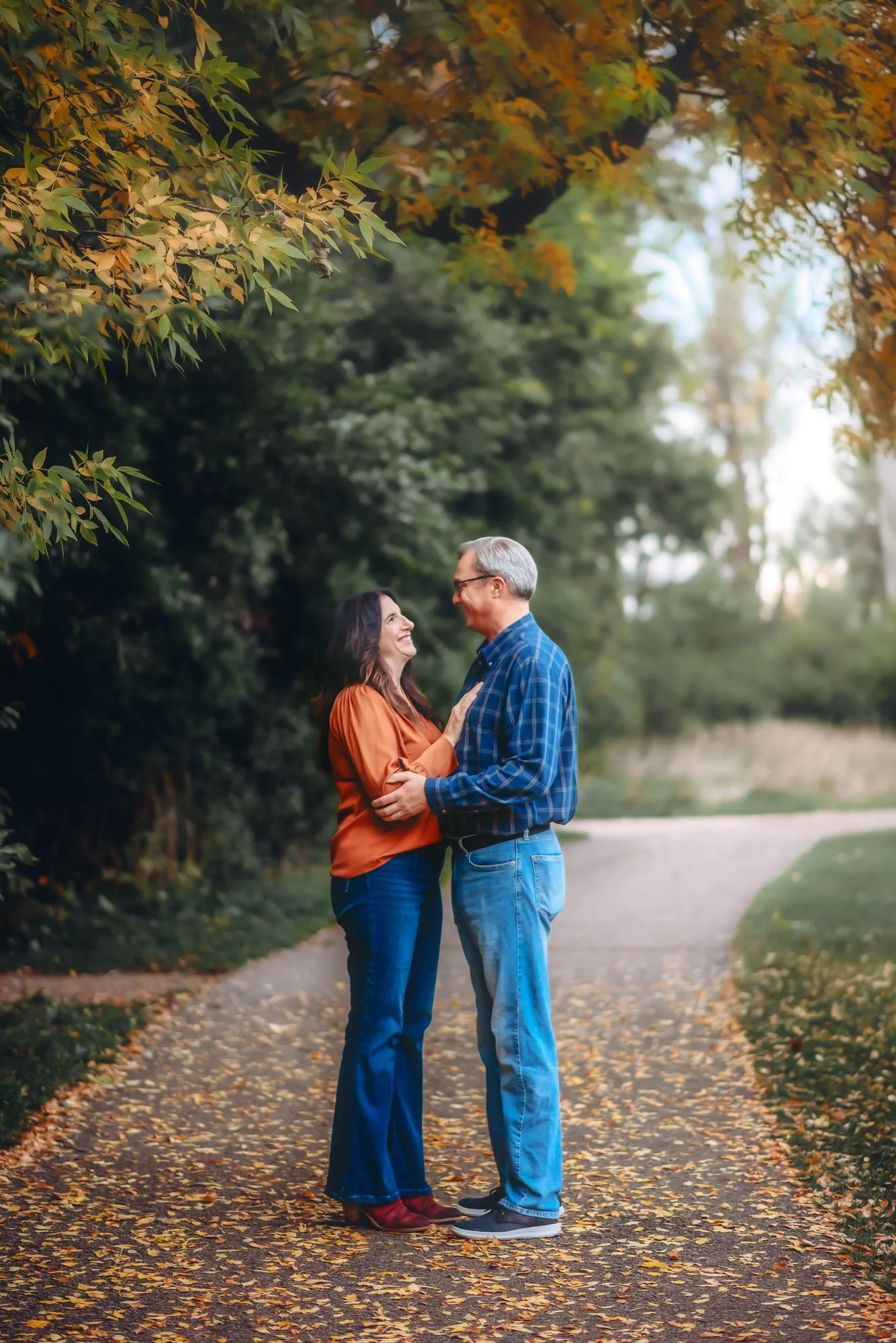 A middle-aged couple stands close together on a fall pathway, smiling at each other, with autumn foliage overhead.
