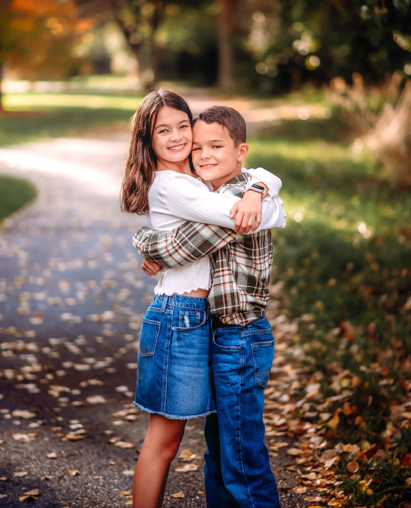 A young girl and boy hugging each other outdoors during autumn in a park with fallen leaves.