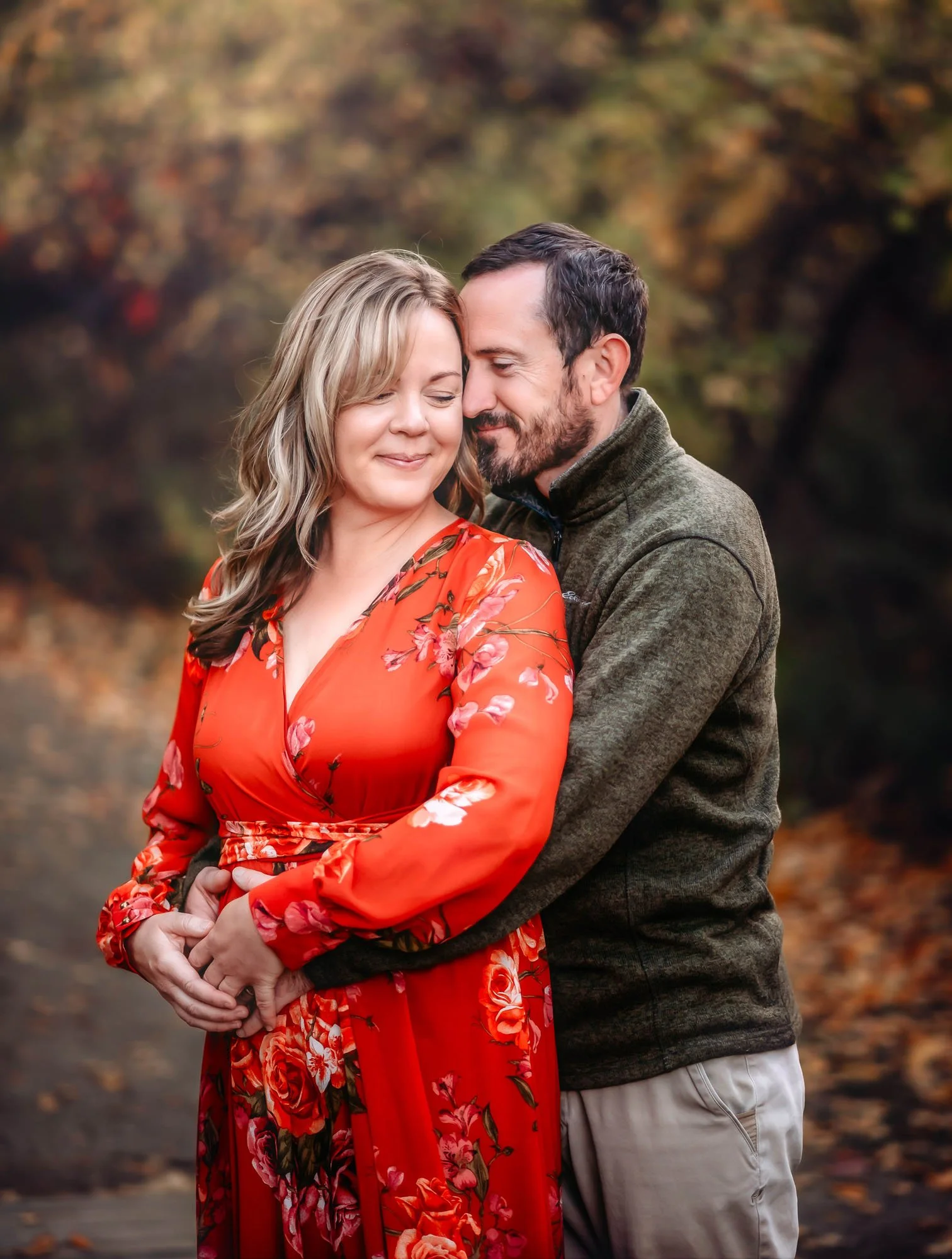 A man and woman are embracing outdoors during fall. The woman is smiling with her eyes closed, wearing a red floral dress, and the man is gently resting his forehead against her temple with a slight smile. The background features autumn trees with co