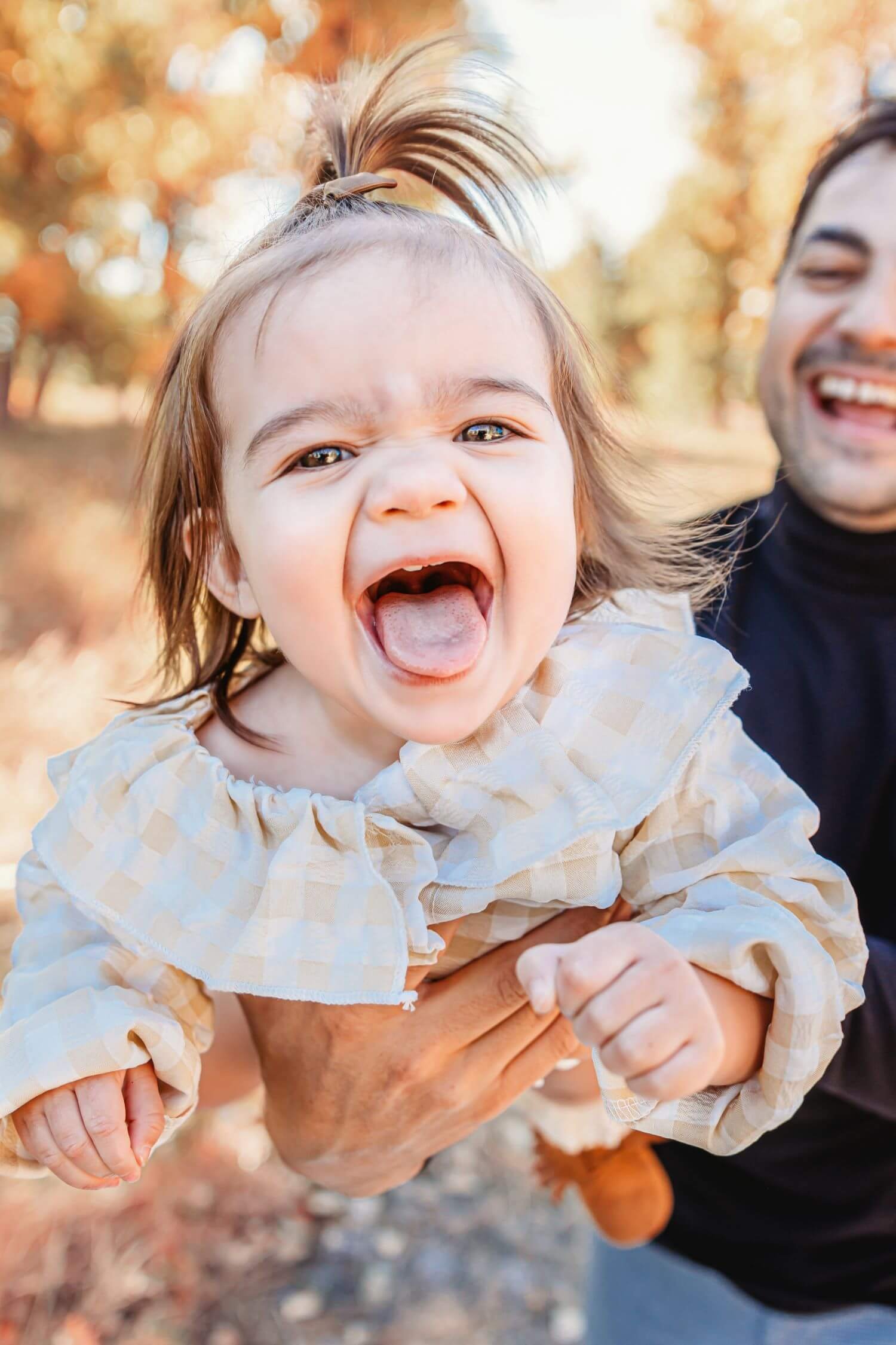 A smiling toddler sticking her tongue out with an adult holding her and a brown dog in the background, outdoors with autumn foliage.