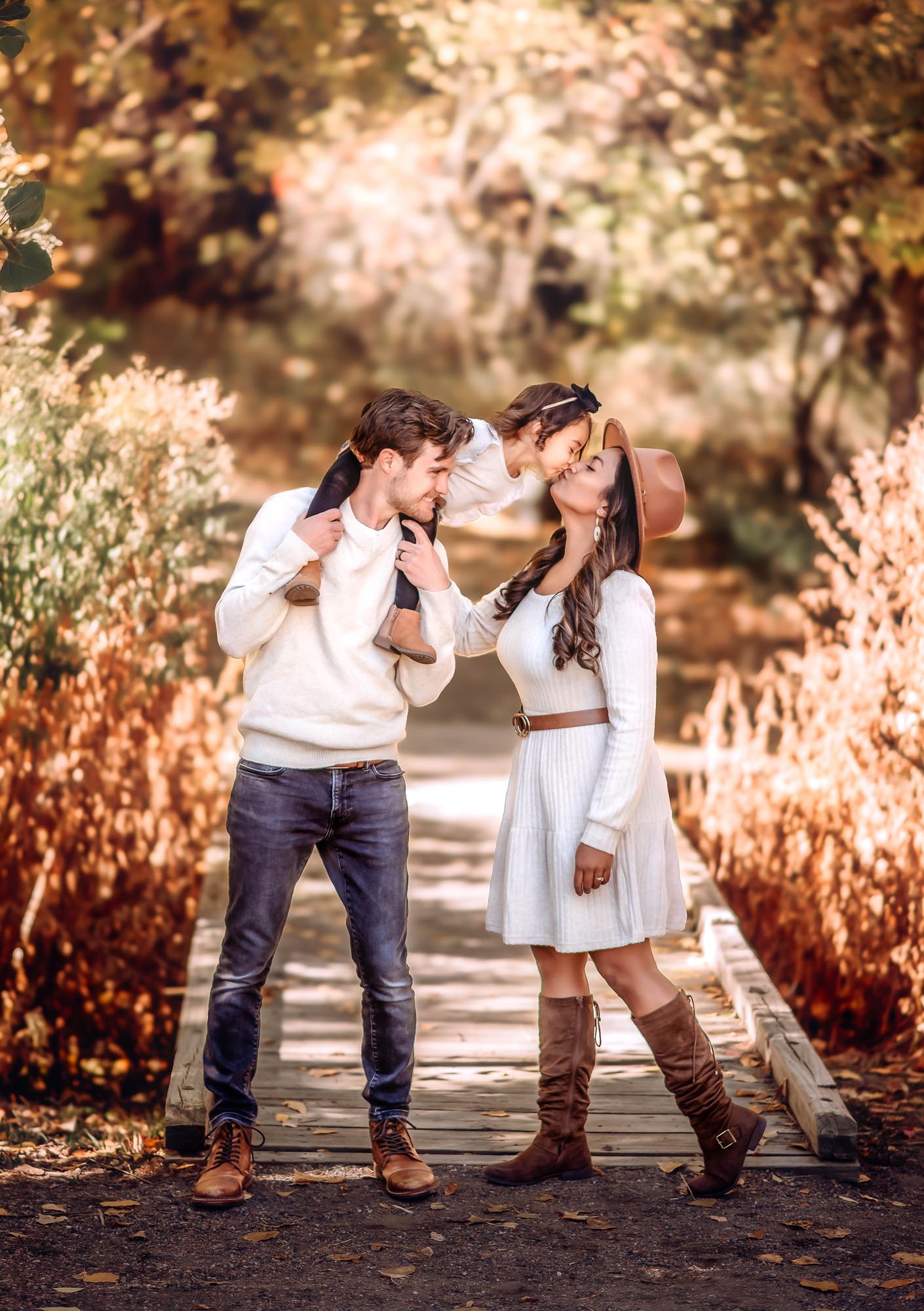 A man, woman, and young girl standing on a wooden bridge surrounded by autumn trees, with the girl leaning over to kiss the woman, who is wearing a hat.