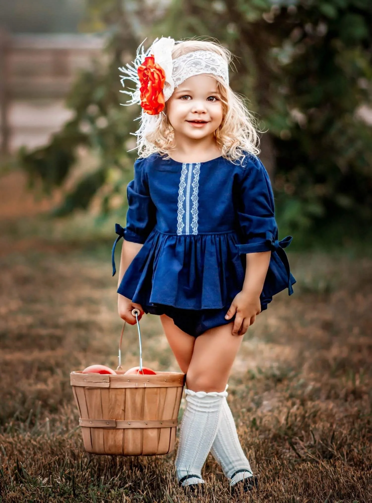 Young girl with curly blonde hair wearing a navy blue dress, white knitted leg warmers, and a lace headband with a large orange flower, holding a basket filled with apples outdoors in a park or garden.