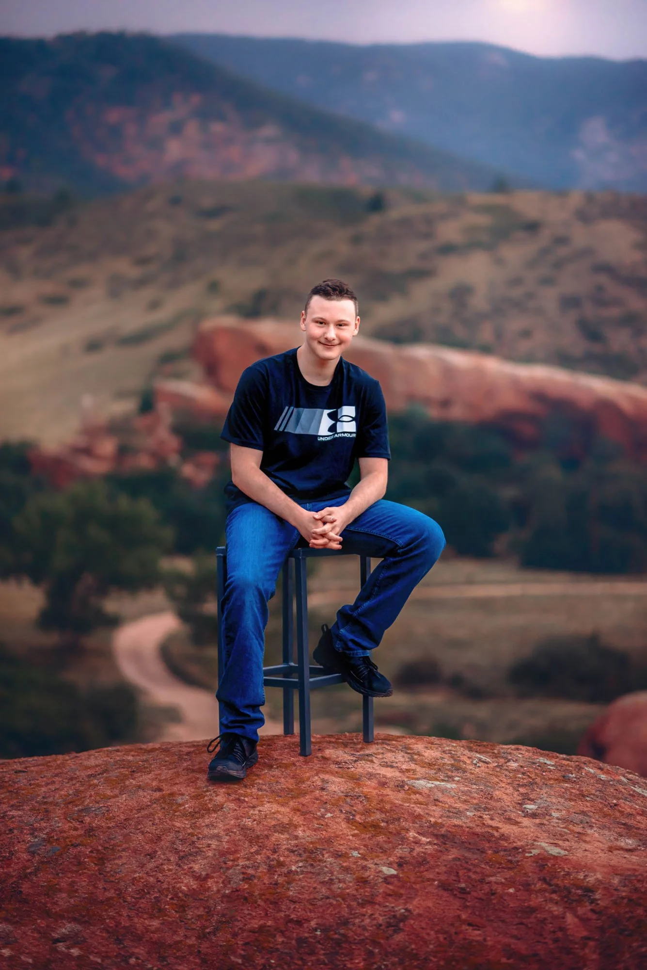 A young man sitting on a tall stool on reddish rock, smiling, with a blurred landscape of hills and a cloudy sky in the background.