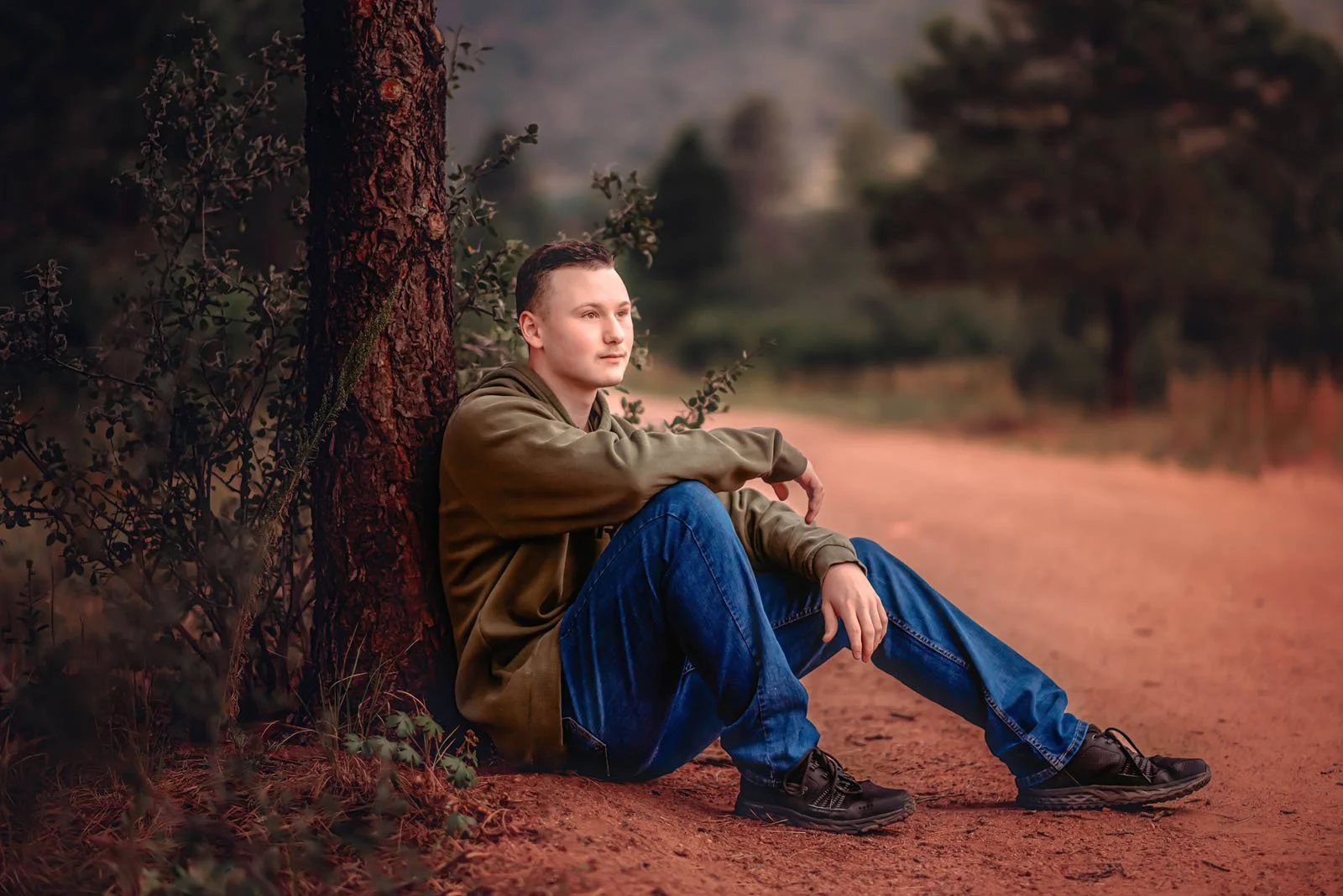 A young man sitting on the ground leaning against a tree in a natural outdoor setting, with a dirt path and trees in the background during sunset or early dusk.
