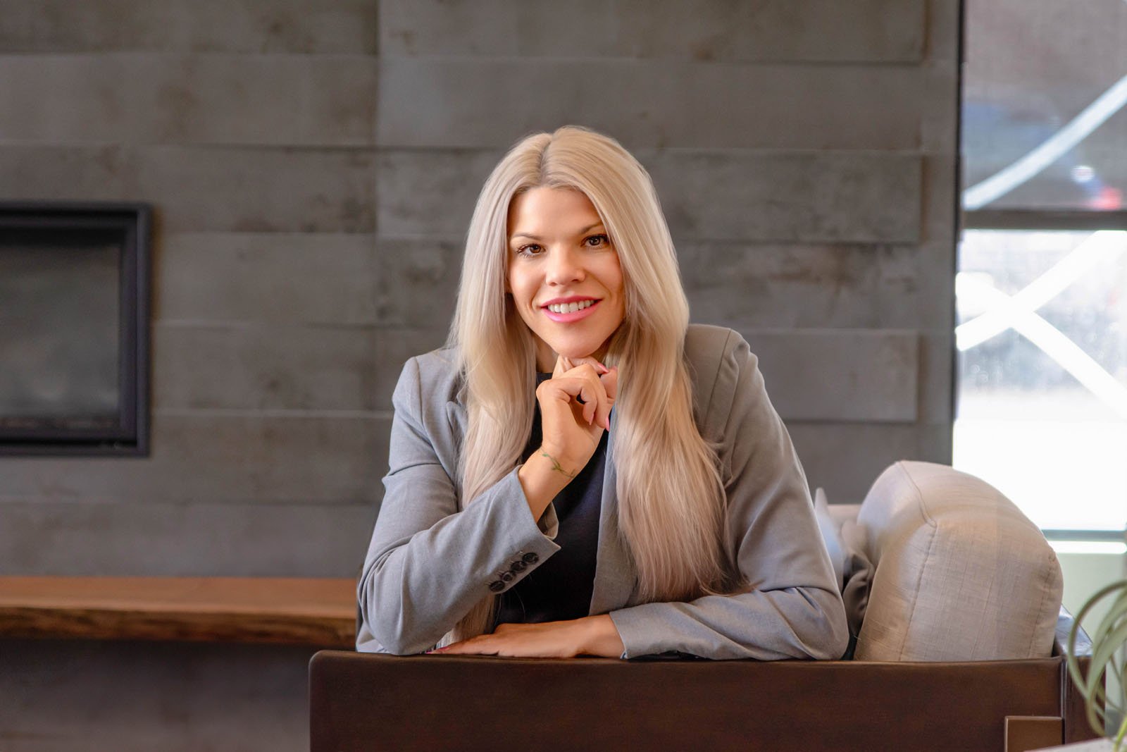 A woman with long blonde hair wearing a grey blazer sitting at a table in a modern indoor setting with a dark wood wall and large windows behind her.