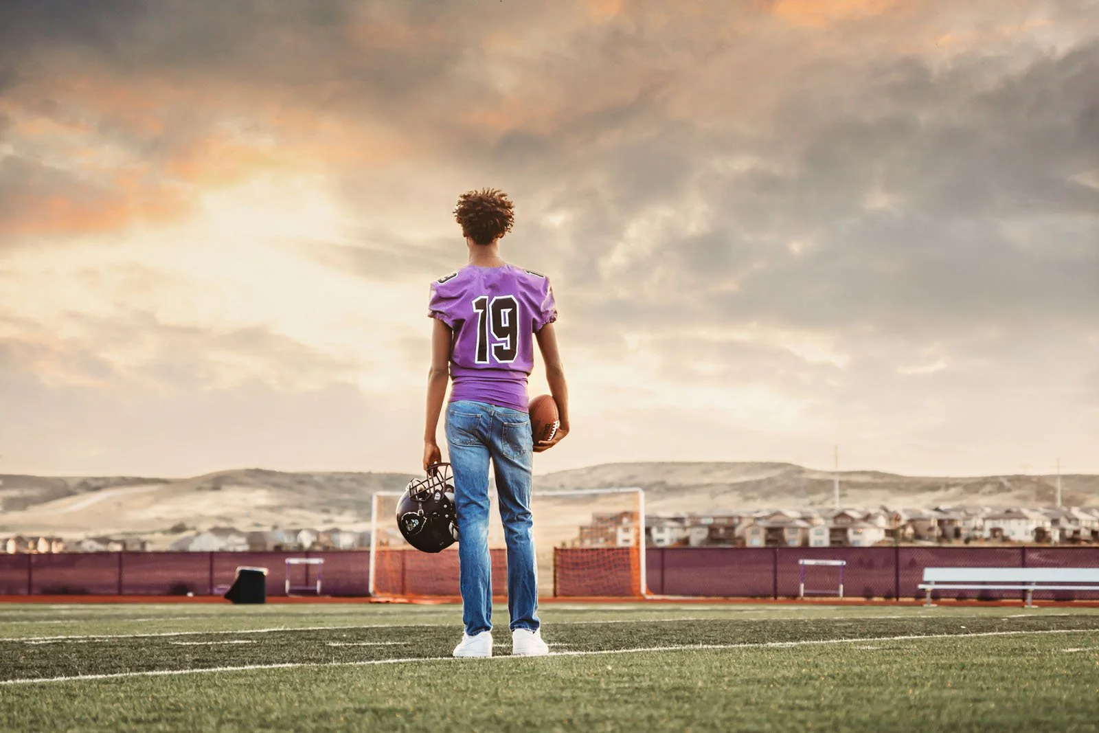 A young male football player standing on a field at sunset, facing away from the camera, holding a football and a helmet, with goalposts and a residential area in the background.