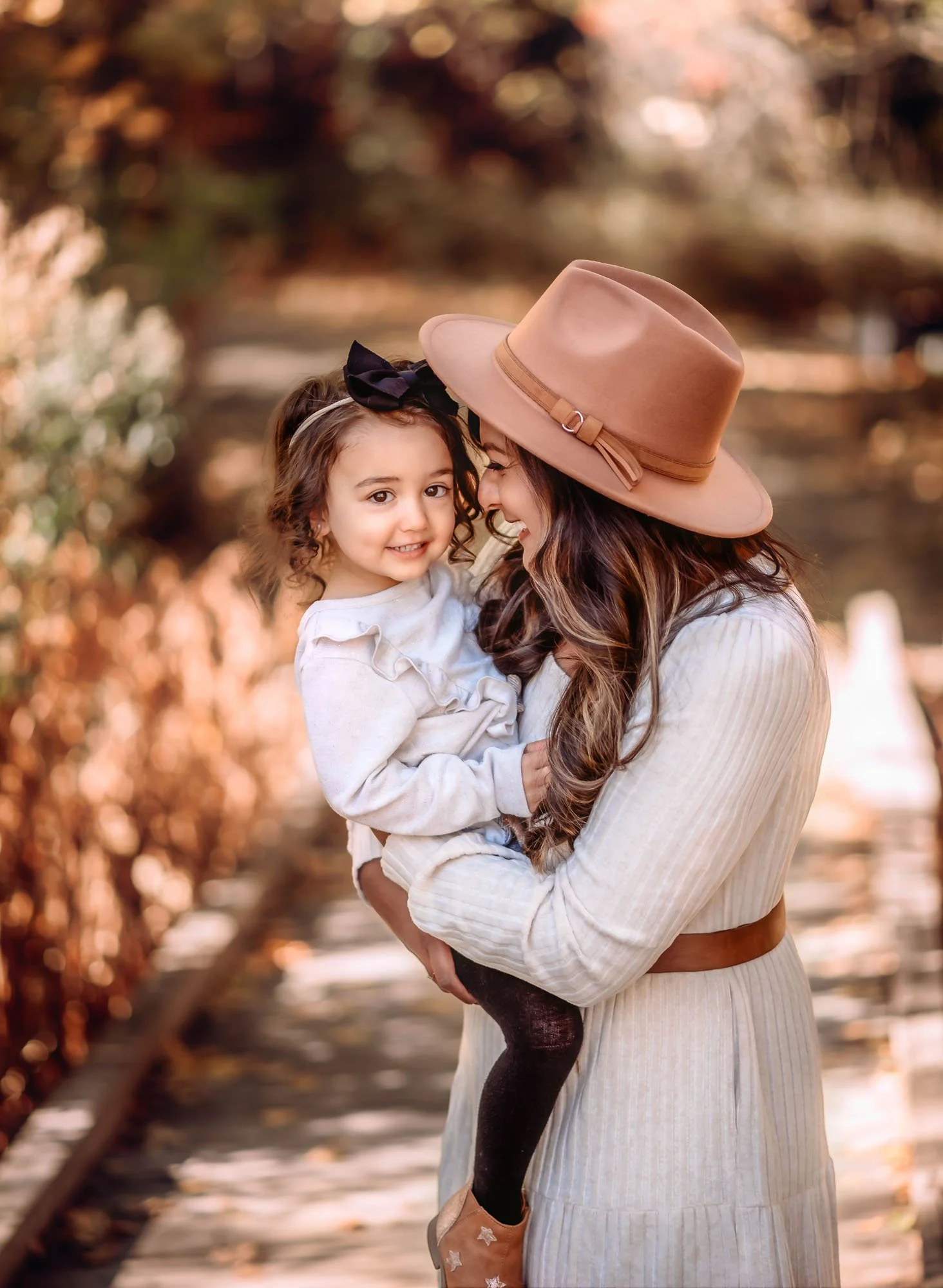 A woman wearing a beige hat with a black bow and a light beige dress holding a young girl with curly hair and a black headband, by a railroad track with autumn leaves in the background.