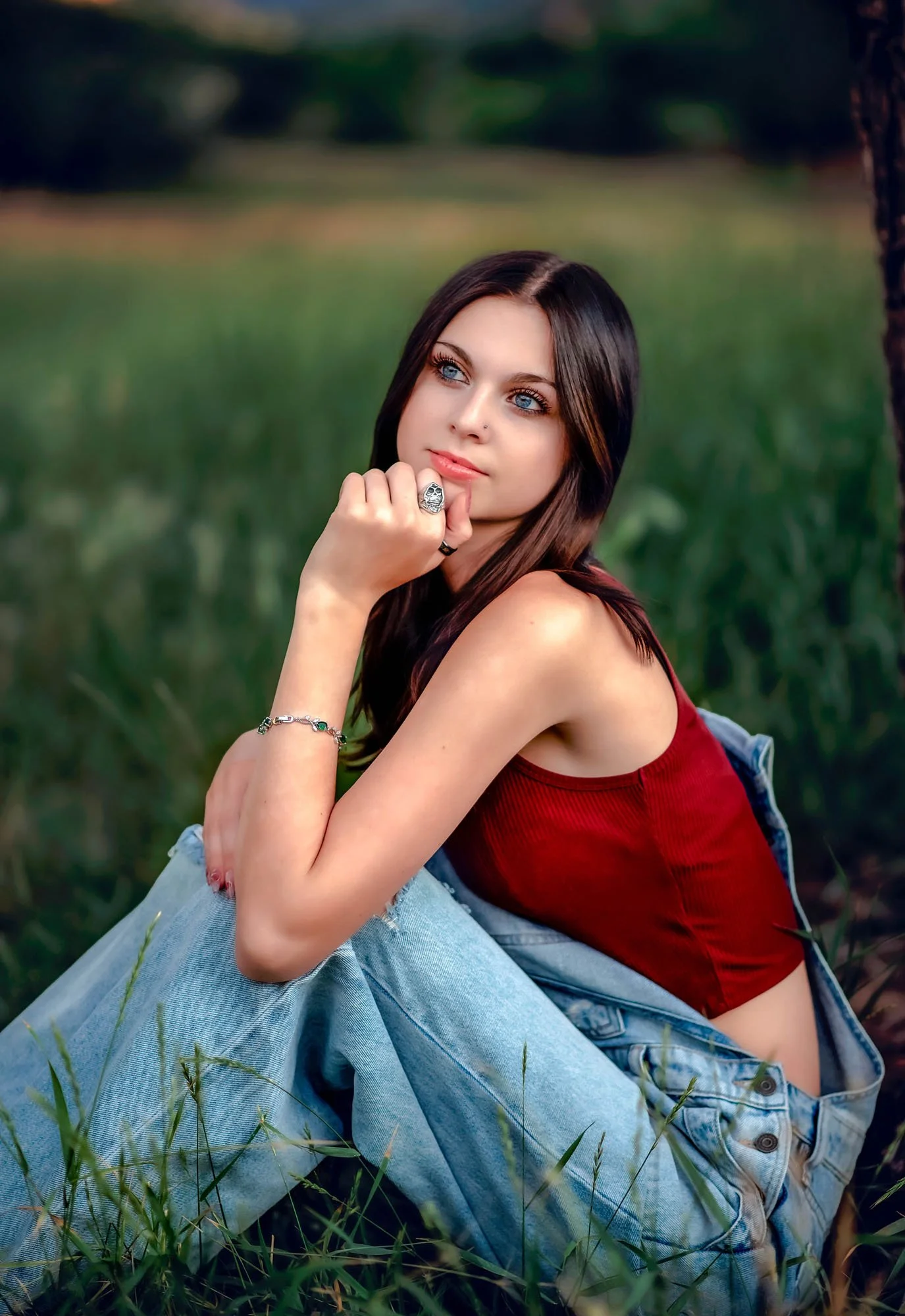 A young woman with blue eyes and dark brown hair sitting on grass outdoors, wearing a red top, blue jeans, and a denim jacket, resting her chin on her hand with a thoughtful expression.