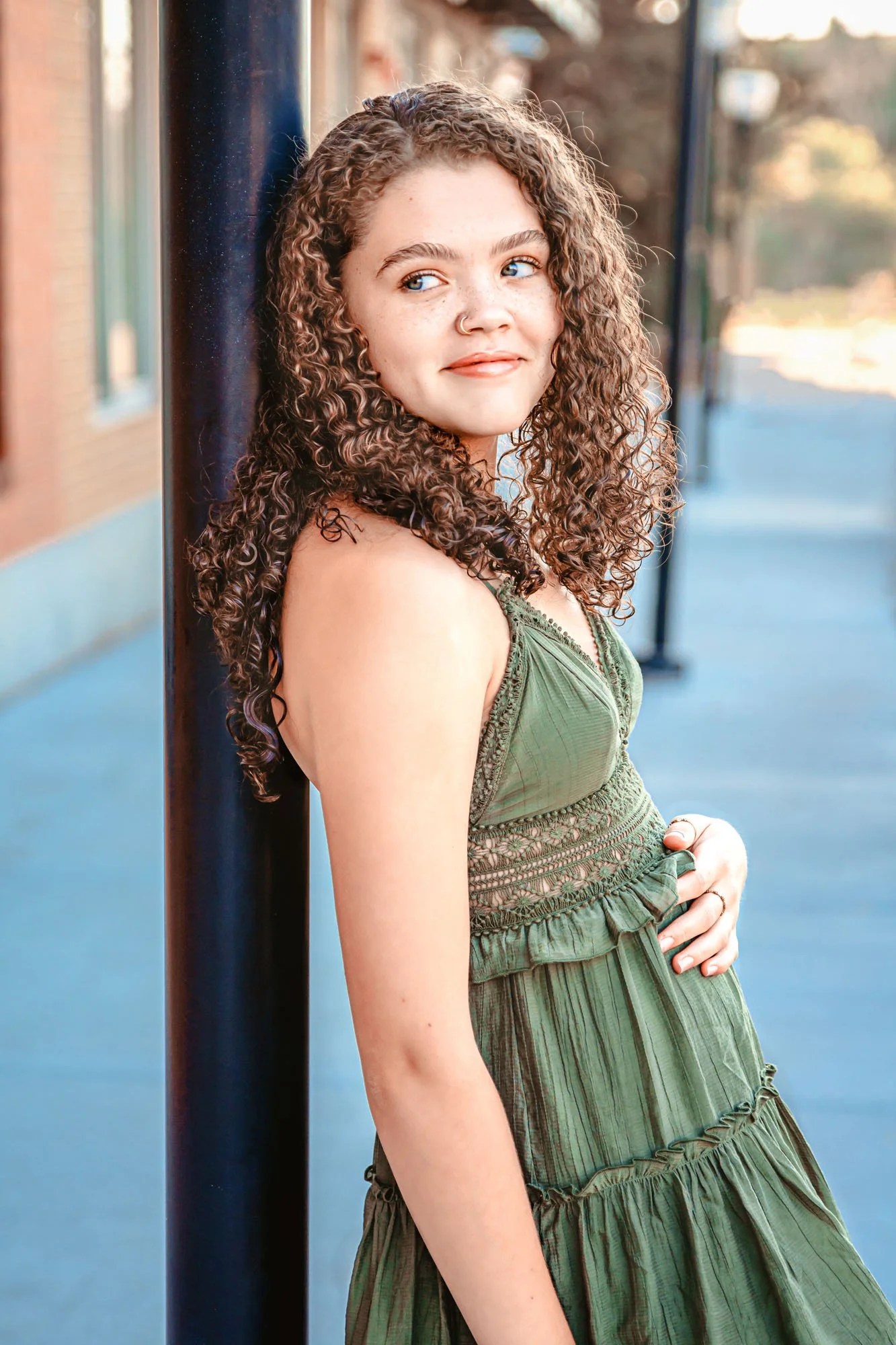 A young woman with curly hair leaning against a black pole on a sidewalk, wearing a green dress, with blurred trees and buildings in the background.
