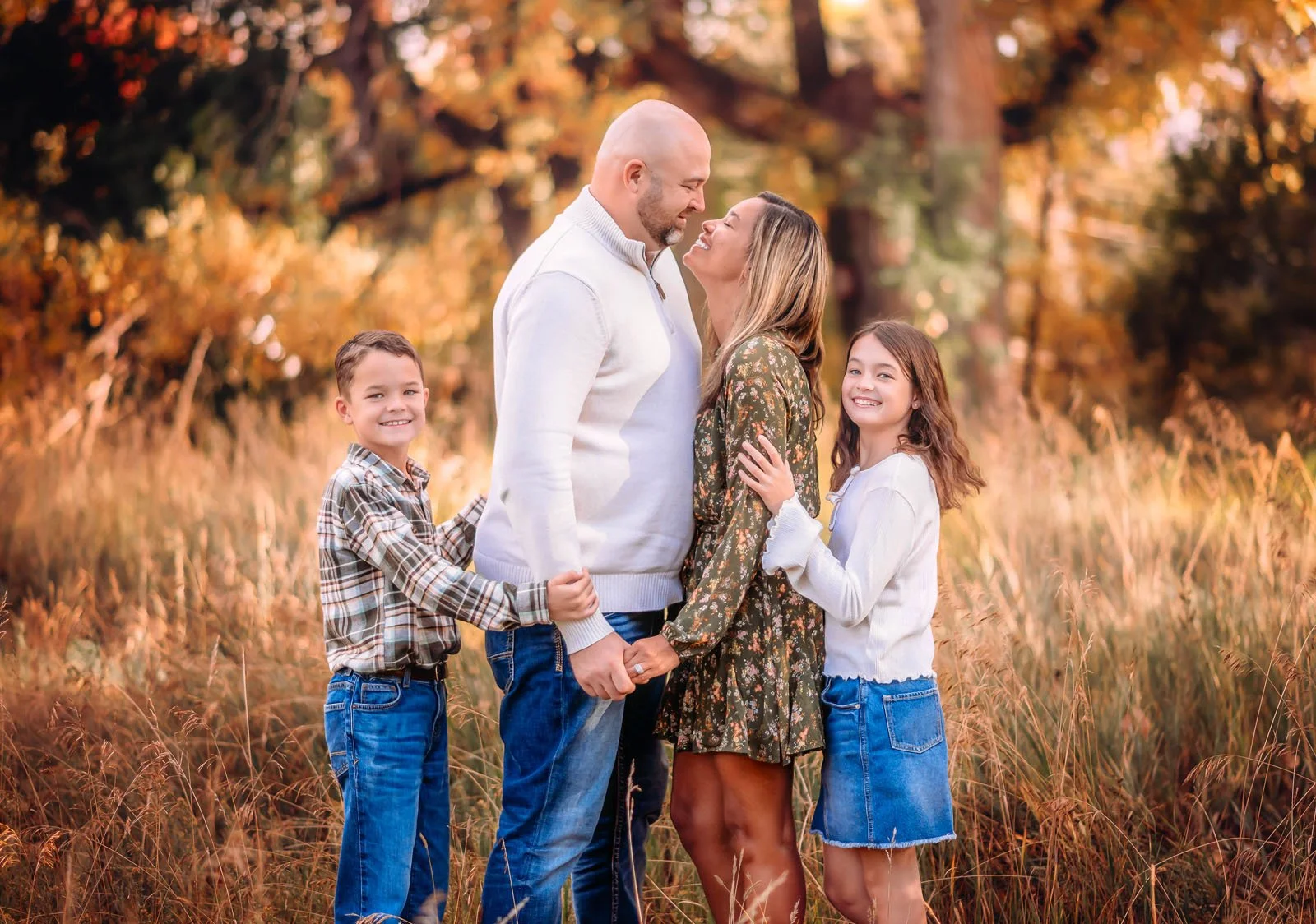 A family of five, including two children and two adults, holding hands and looking at each other affectionately in a field during autumn.