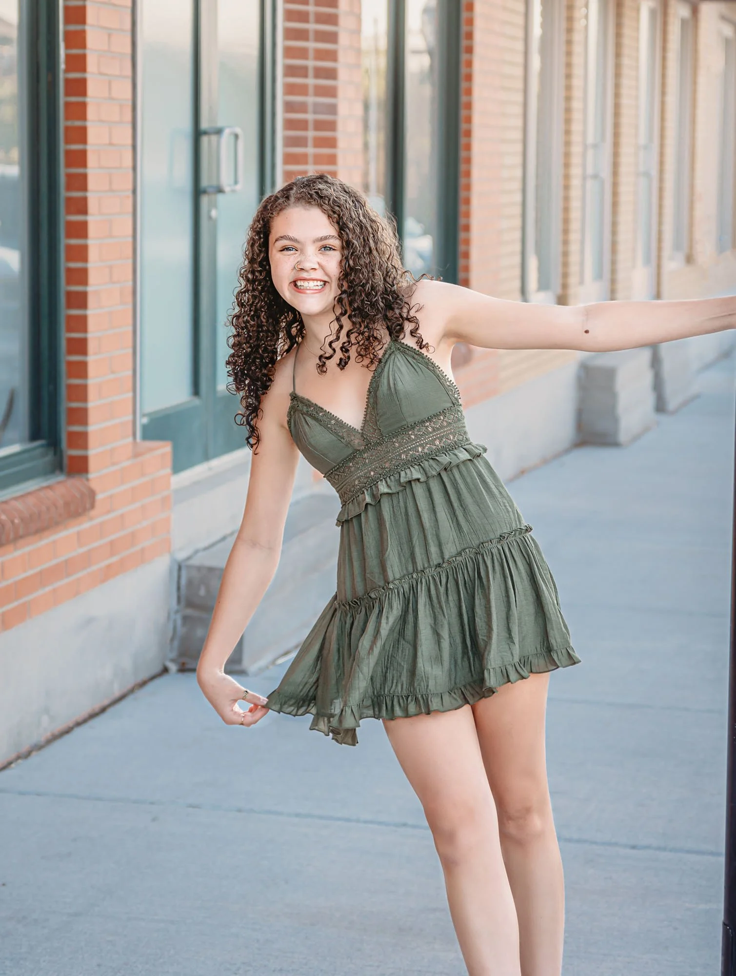A young woman with curly brown hair, smiling and wearing a green dress, extending her right arm and holding the hem of her dress with her left hand, standing on a sidewalk in front of a brick building with large glass windows.