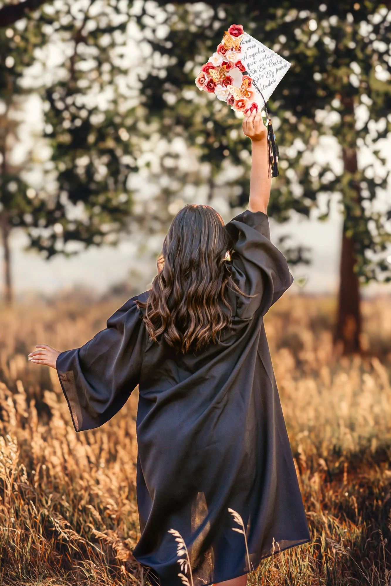 A woman in a black gown holding a decorated graduation cap with flowers and butterflies, celebrating outdoors in a field during sunset.