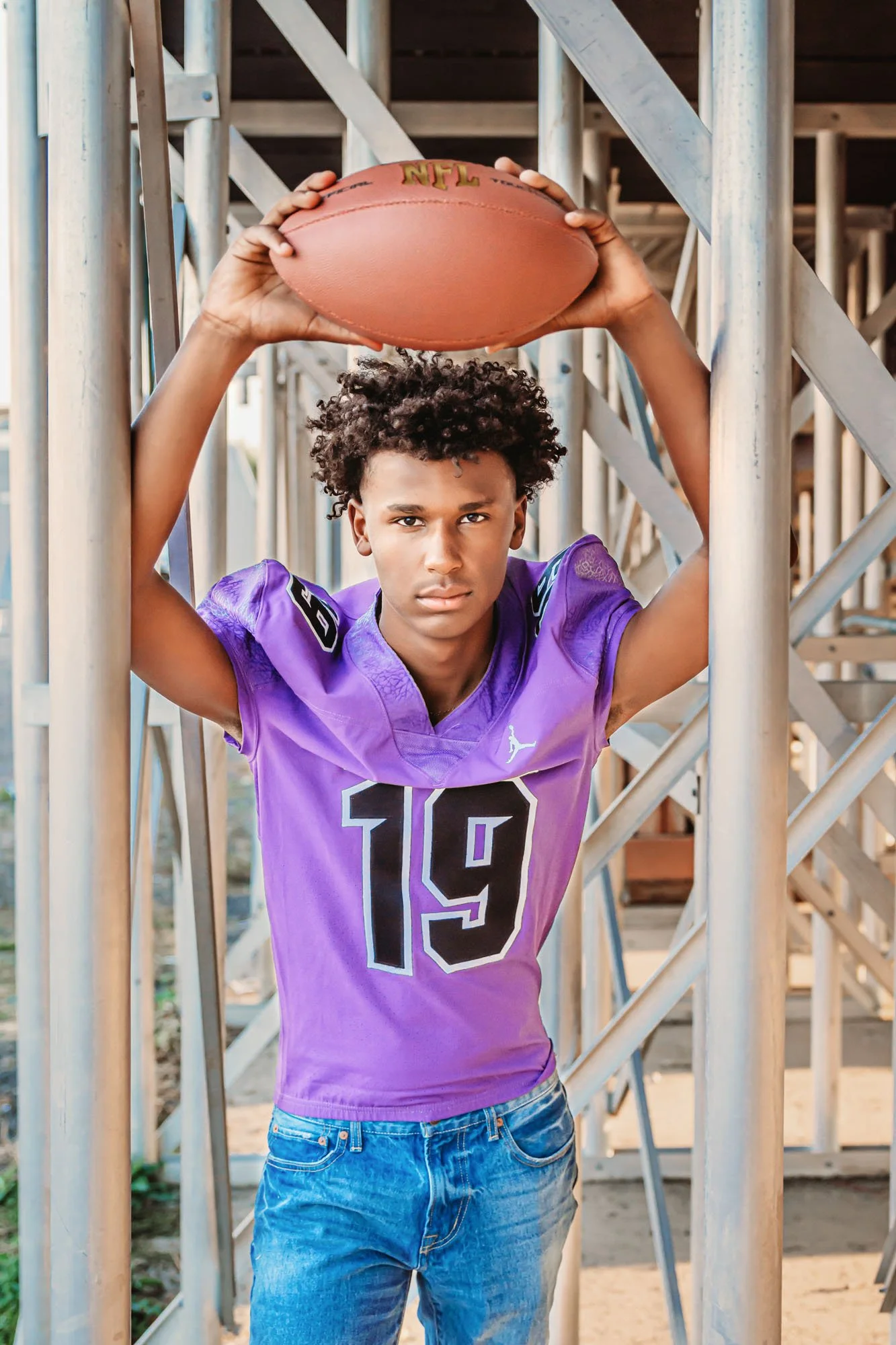 Young man with curly hair wearing a purple sports jersey holding a football above his head, standing amid metal scaffolding.