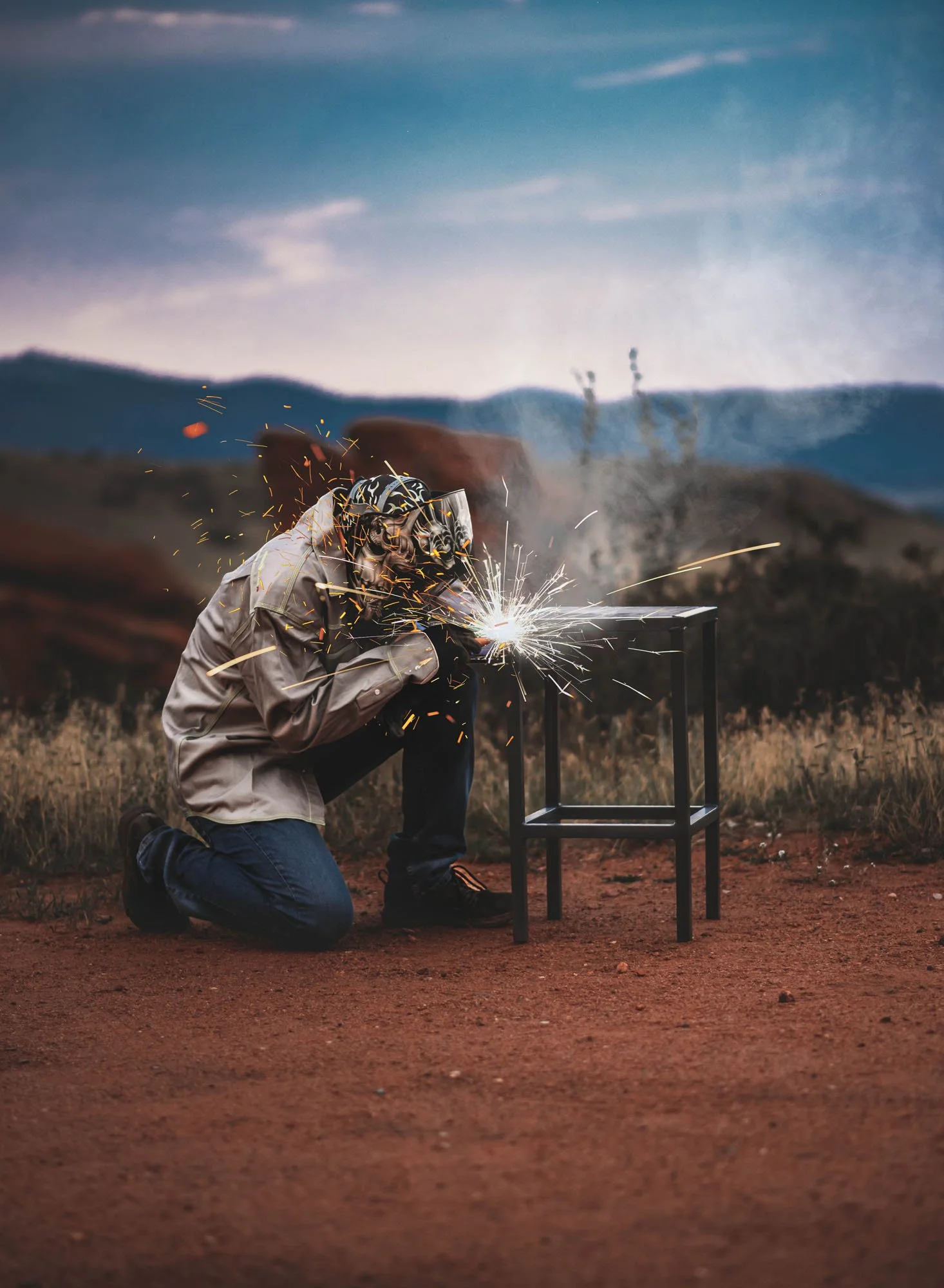 A person welding while kneeling on the ground, sparks flying, outdoors with mountains in the background.