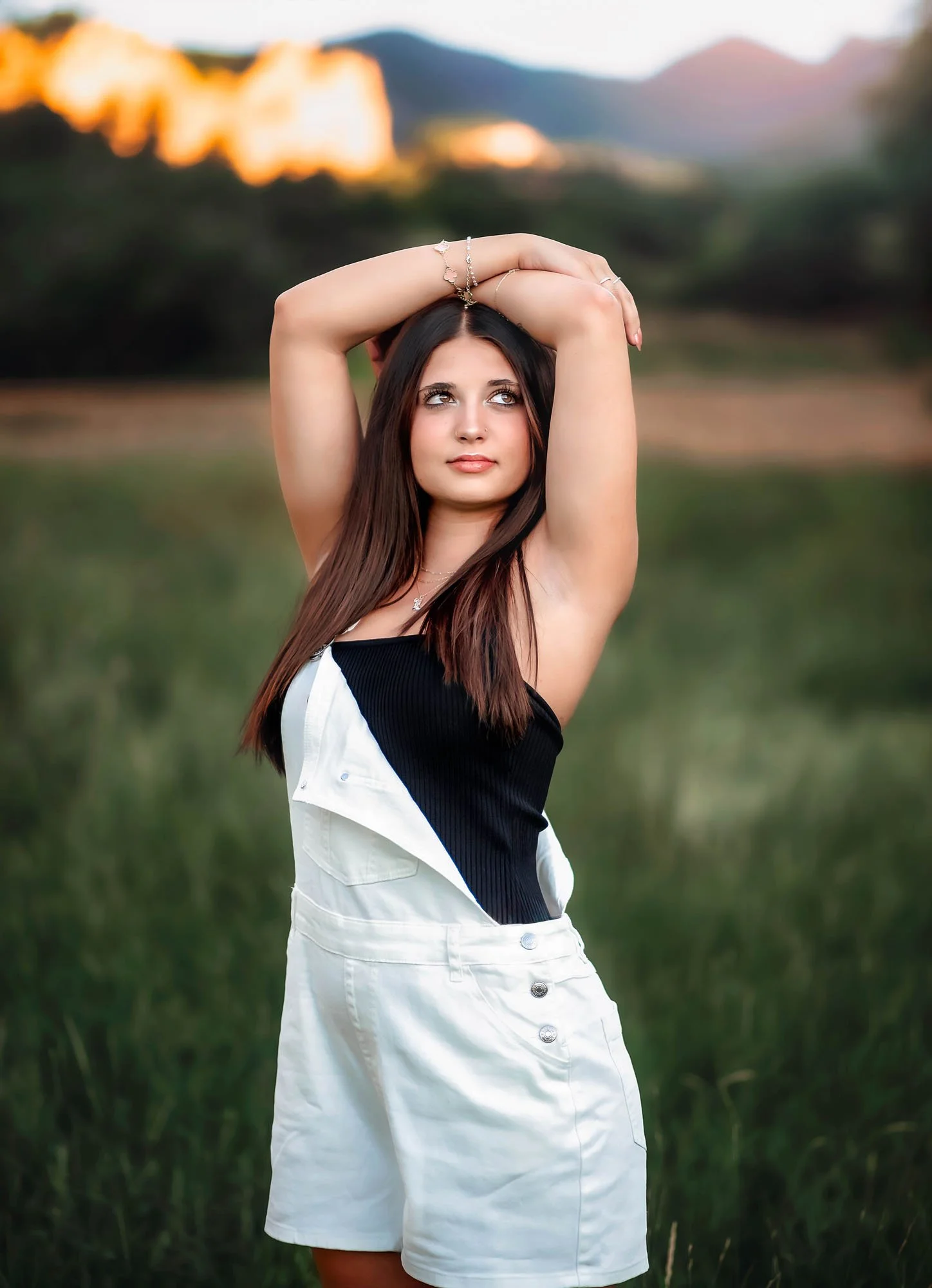 A young woman with long dark hair poses outdoors in a field with tall grass, wearing a black and white top and white high-waisted shorts, with her arms raised above her head and a serene expression.